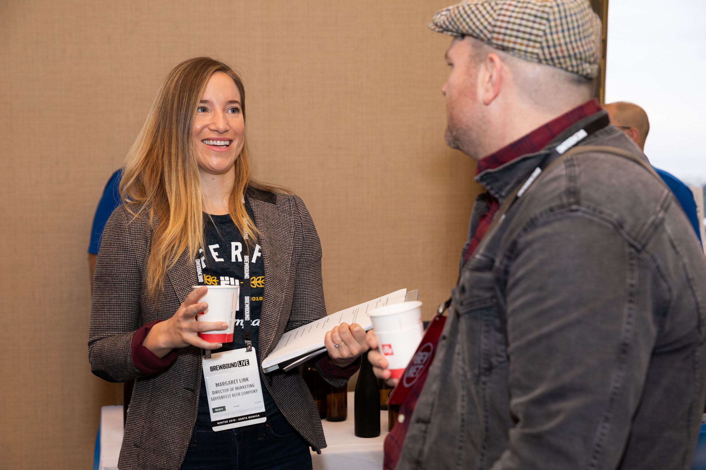 Woman in a tweed blazer holding a coffee cup talking with a man in a flat cap and denim jacket.