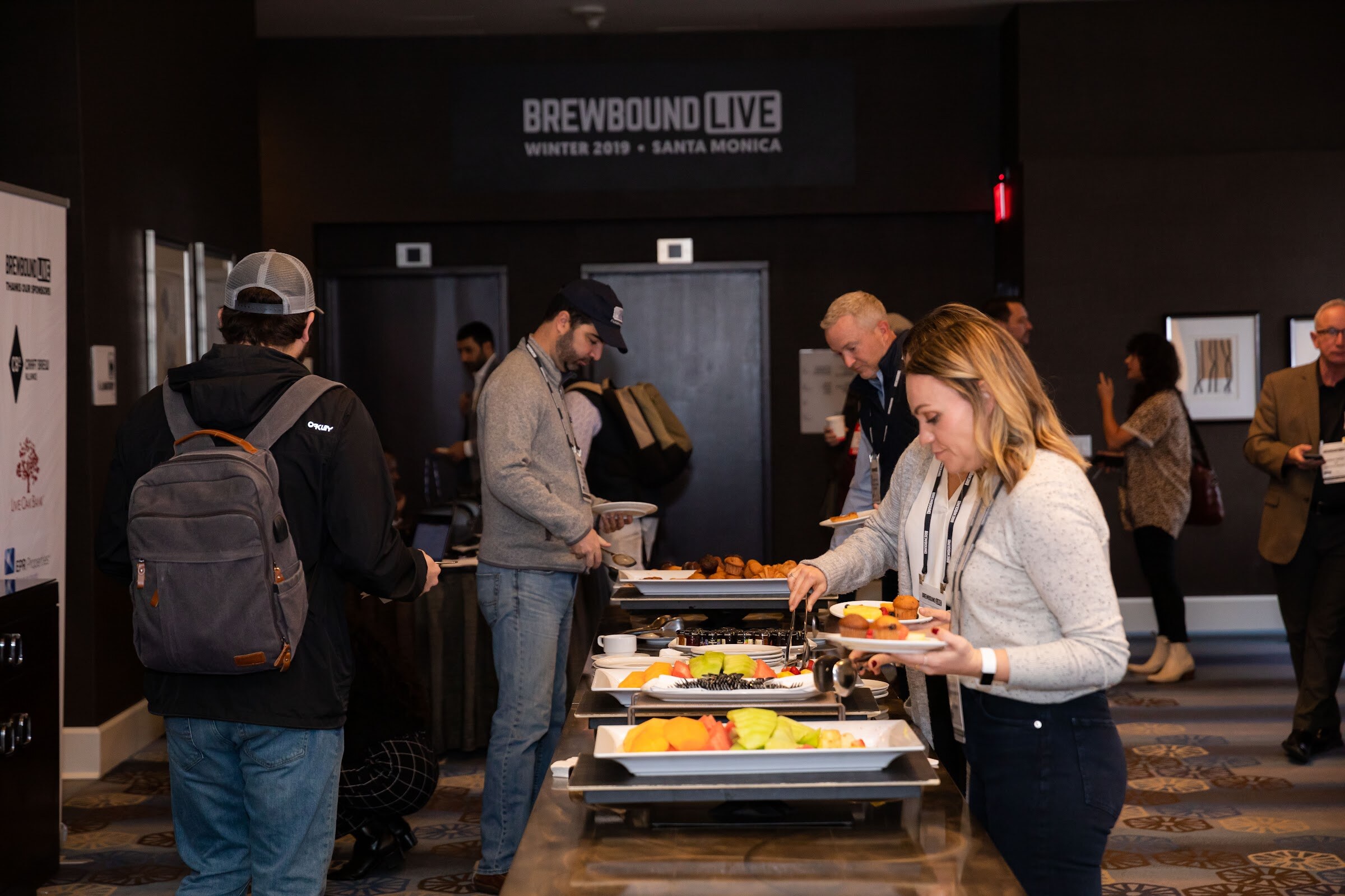 Attendees at a buffet table with fruit and pastries in front of a sign reading Brewbound Live Winter 2019 Santa Monica.