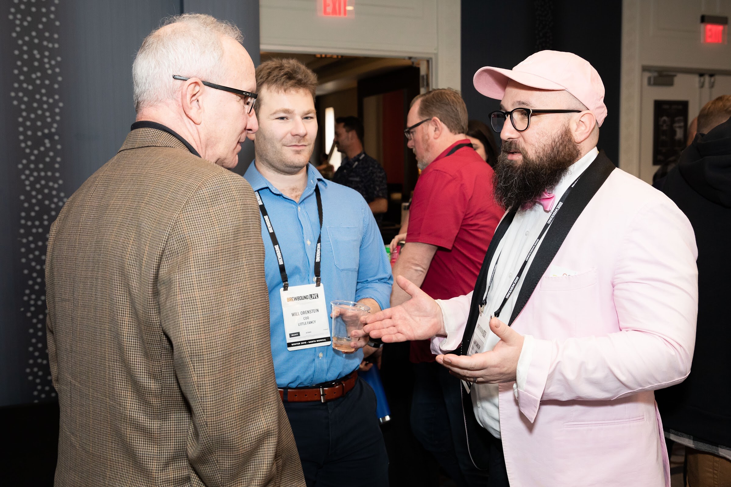 Man in a pink blazer and bow tie speaking with an older man in a tweed jacket and a younger man with a badge.