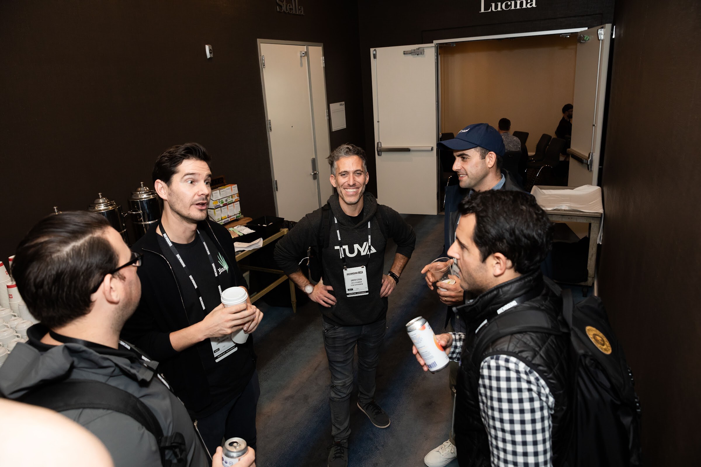 Group of men standing in a circle in a hallway; one wears a TUYA hoodie and others hold cans.