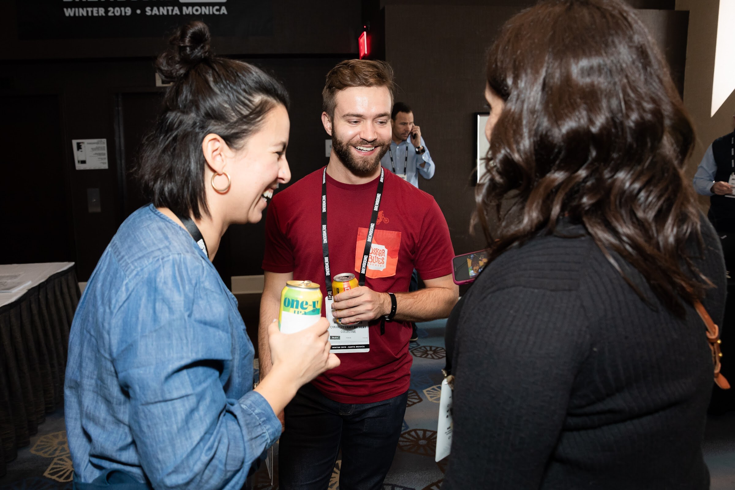 Smiling woman in a denim shirt and a bearded man in a red T-shirt holding cans, with another woman in the foreground.