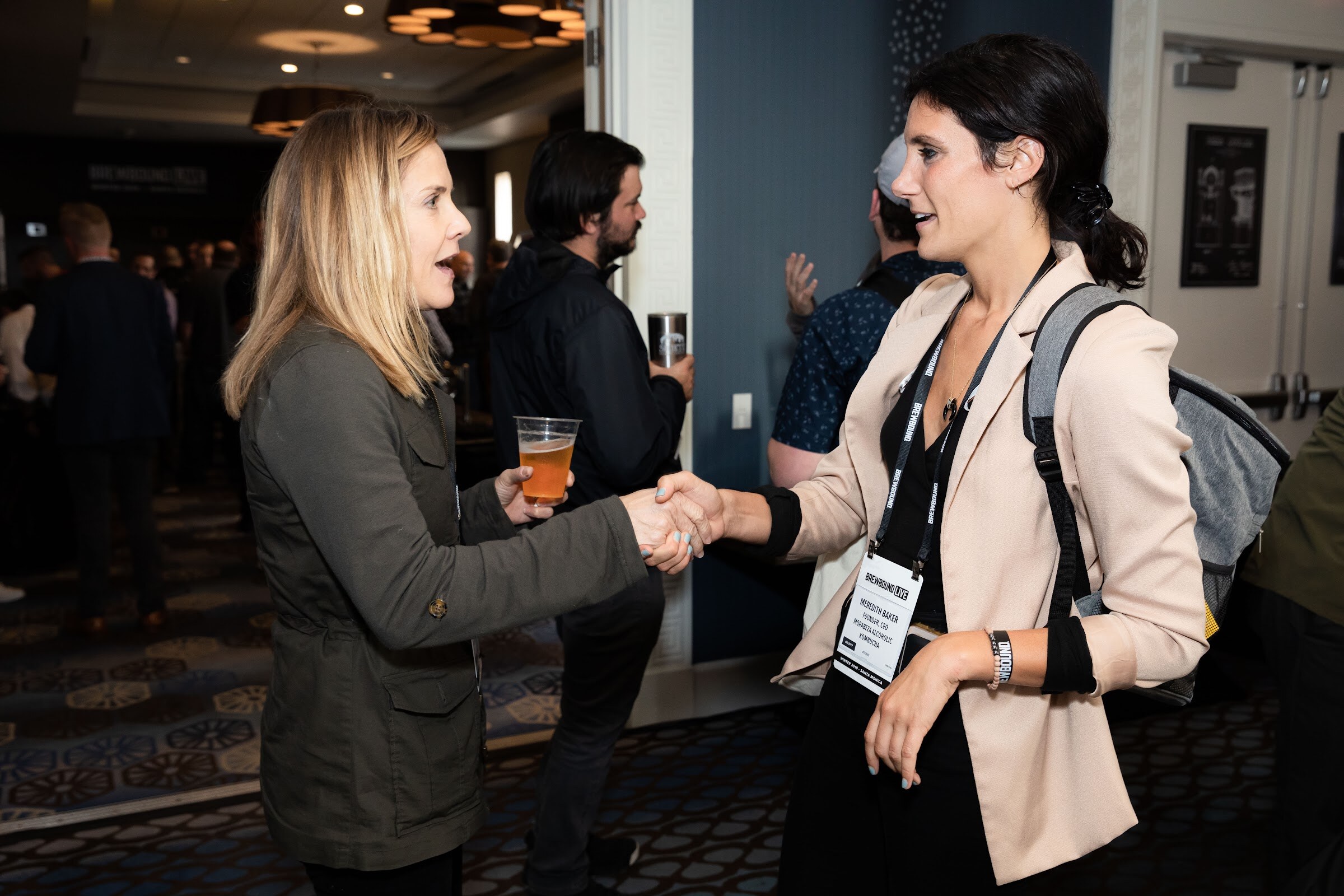Woman in an olive jacket holding a beer shaking hands with a woman in a beige blazer.