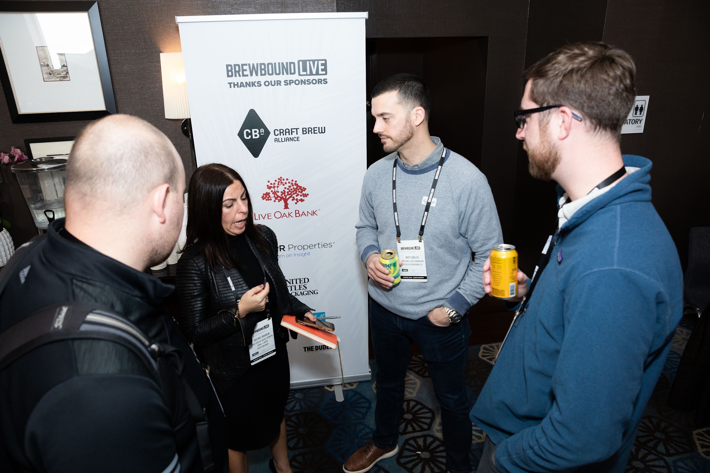 Four people holding cans and talking next to a Brewbound Live Thanks Our Sponsors pop-up banner.