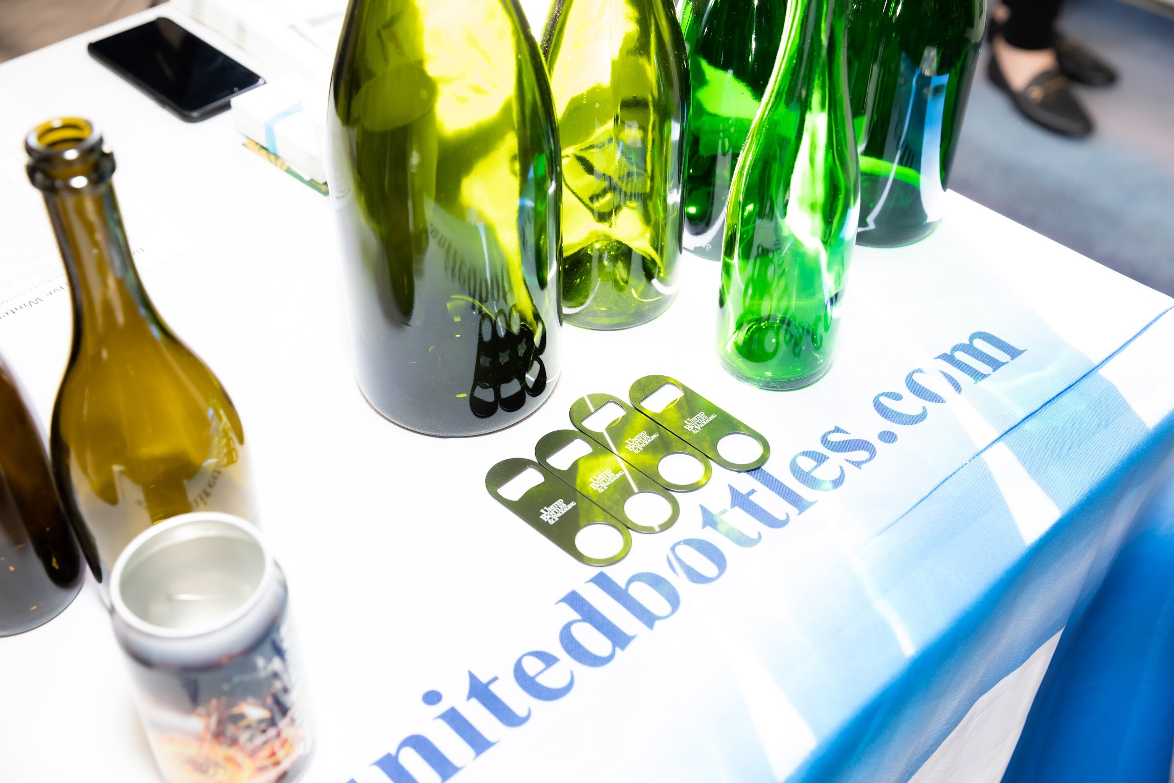 Close-up of green and amber glass bottles and metal bottle openers on a table with a United Bottles and Packaging cloth.