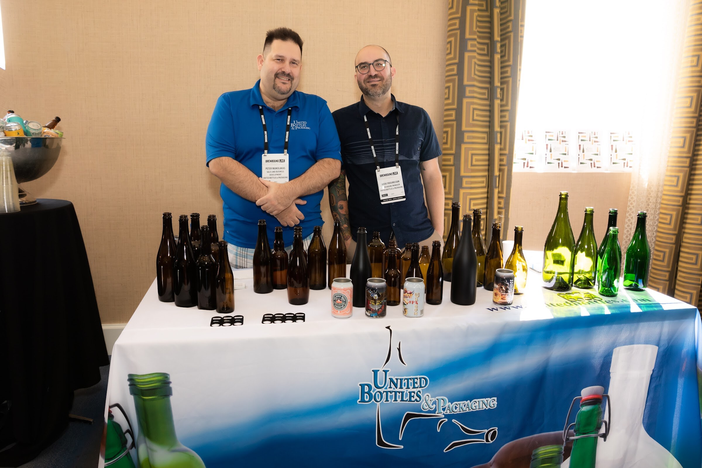 Two men with badges standing behind a table of glass bottles with a United Bottles and Packaging tablecloth.