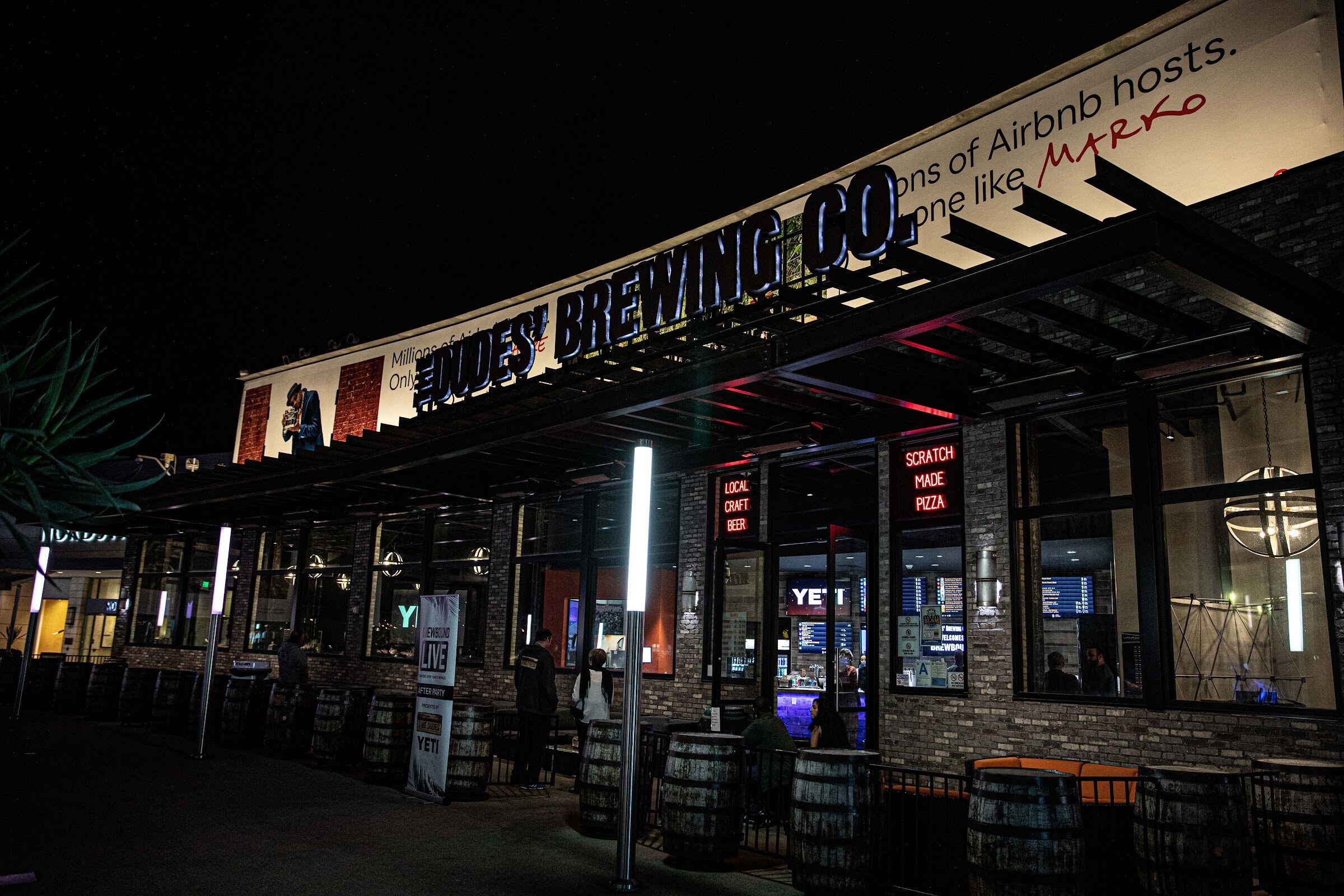 Night view of the Dudes' Brewing Co. storefront with barrel tables outside.