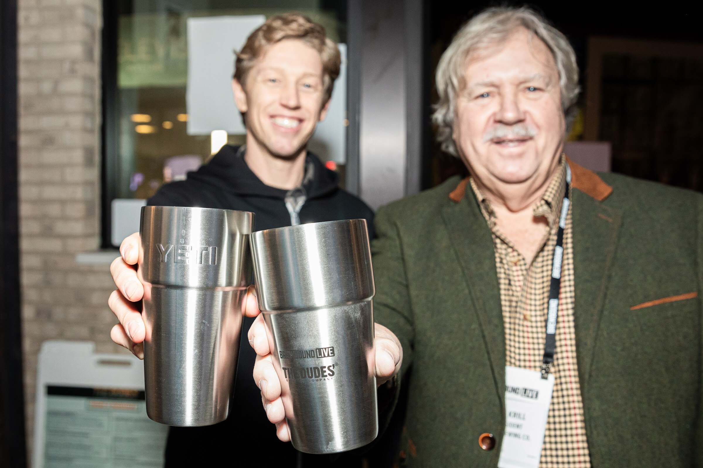Smiling younger man and older man with a lanyard each holding up a stainless steel YETI cup.