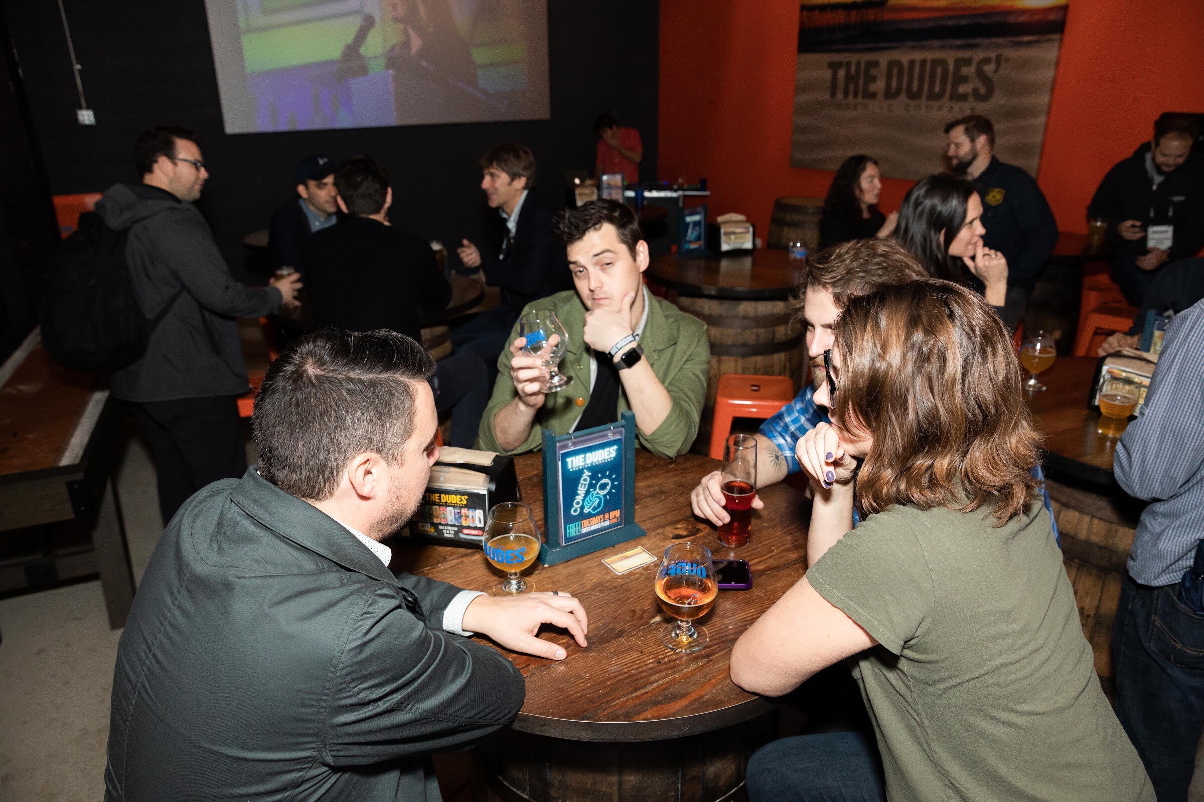 Several guests seated around a barrel-style table with glasses of beer in front of a Dudes' Brewing sign.