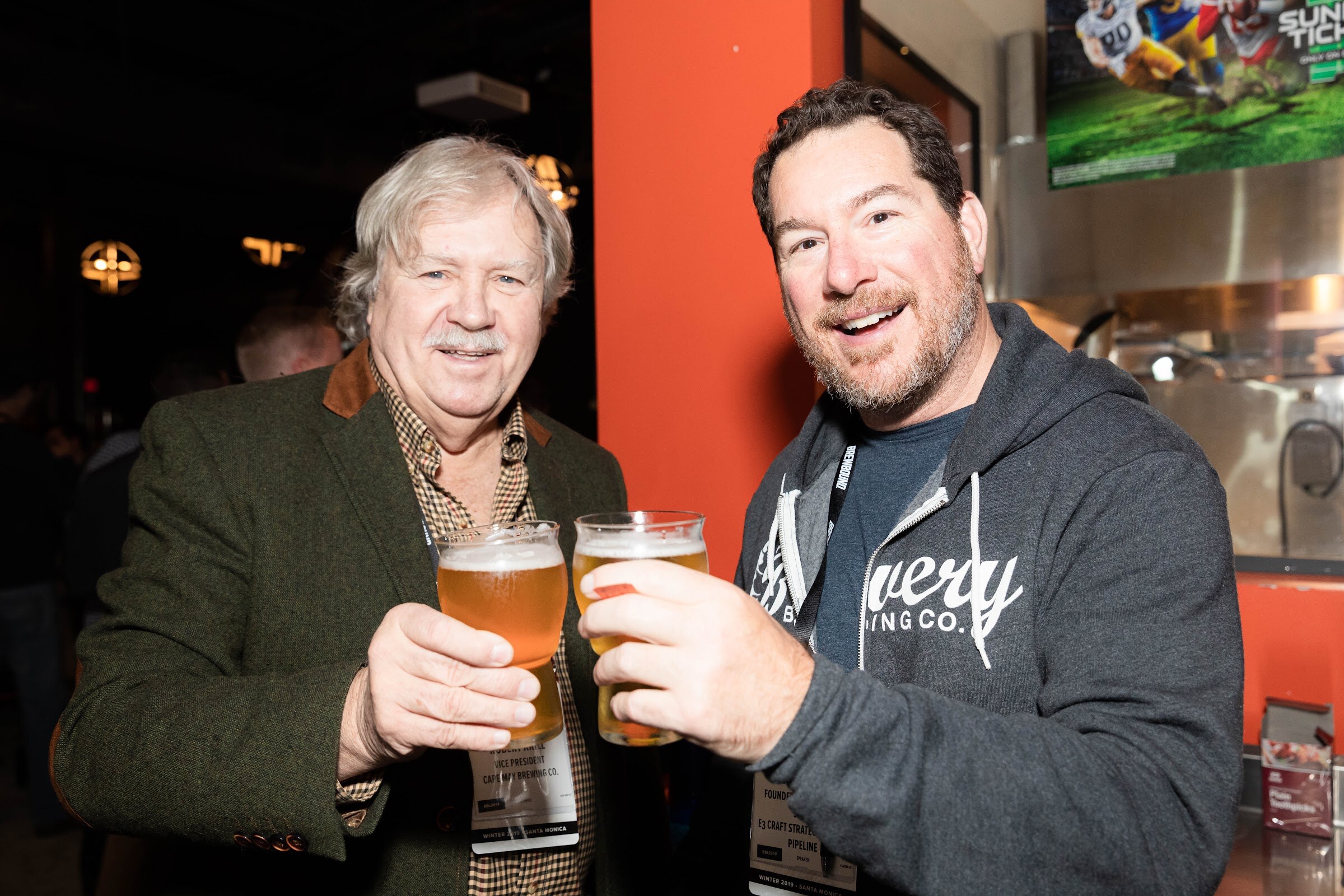 Two smiling men in front of a bar each holding a pint of beer toward the camera.