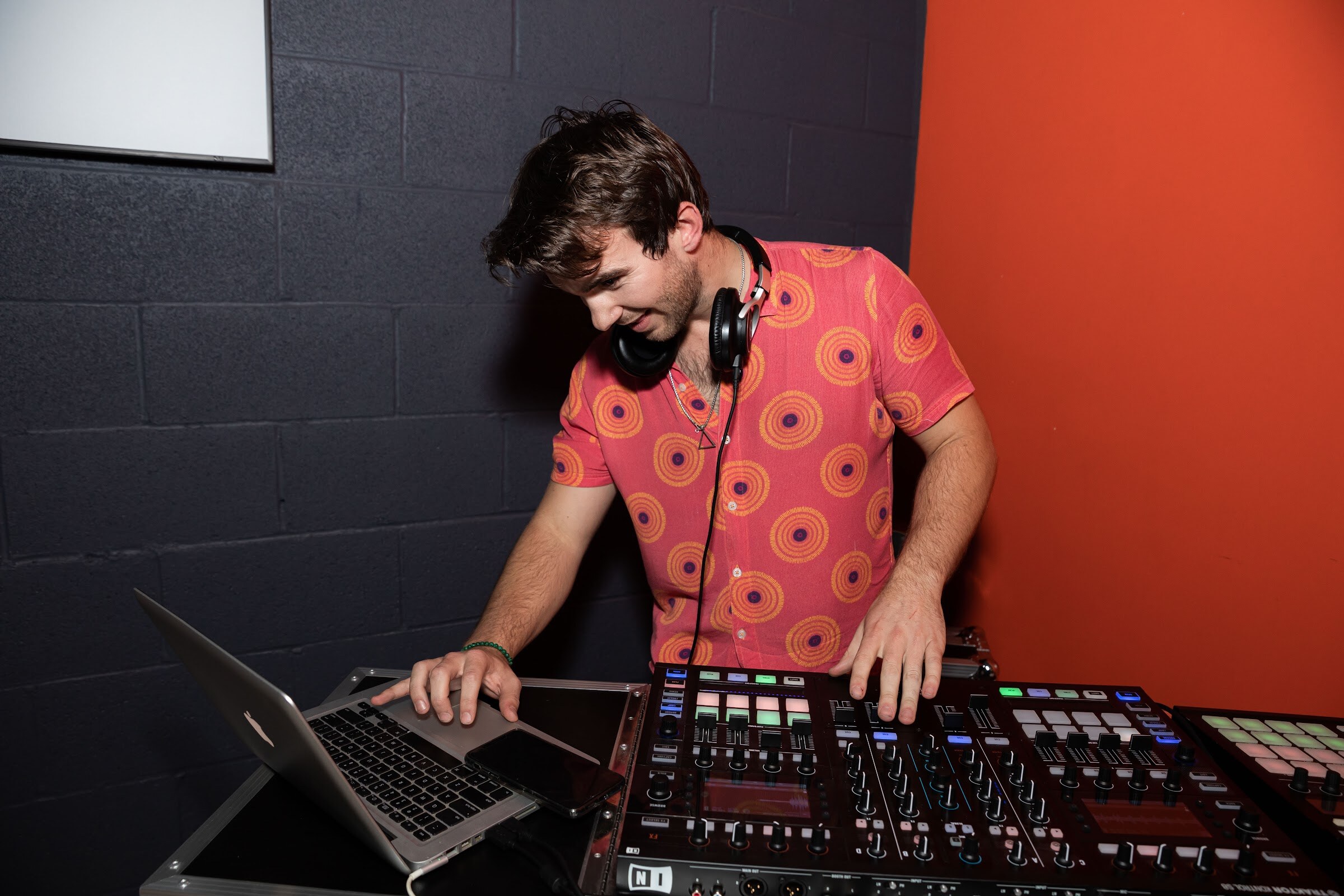 Man in an orange patterned shirt with headphones around his neck at a laptop and DJ controller.