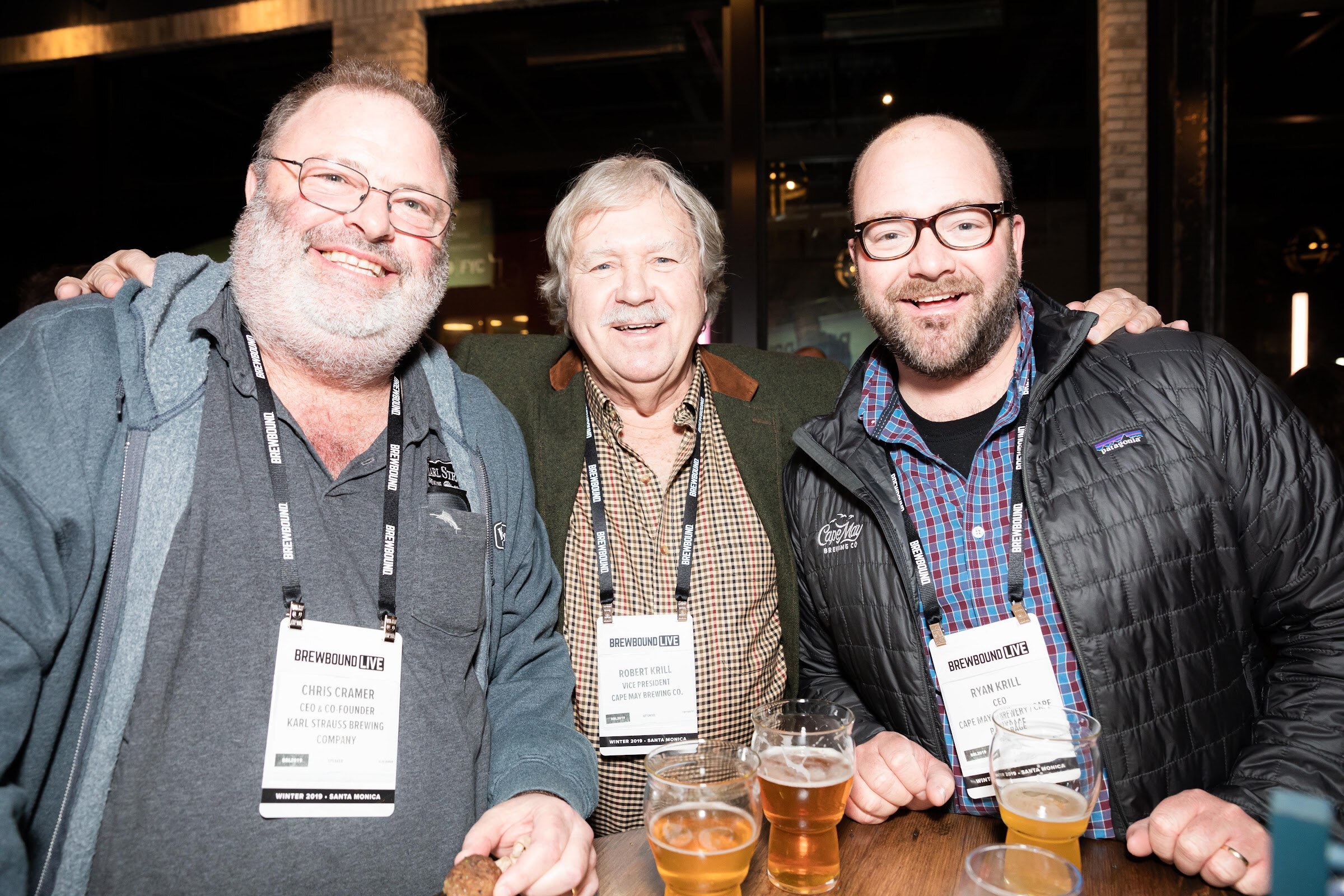 Three smiling men with arms around each other holding glasses of beer, badges on lanyards.