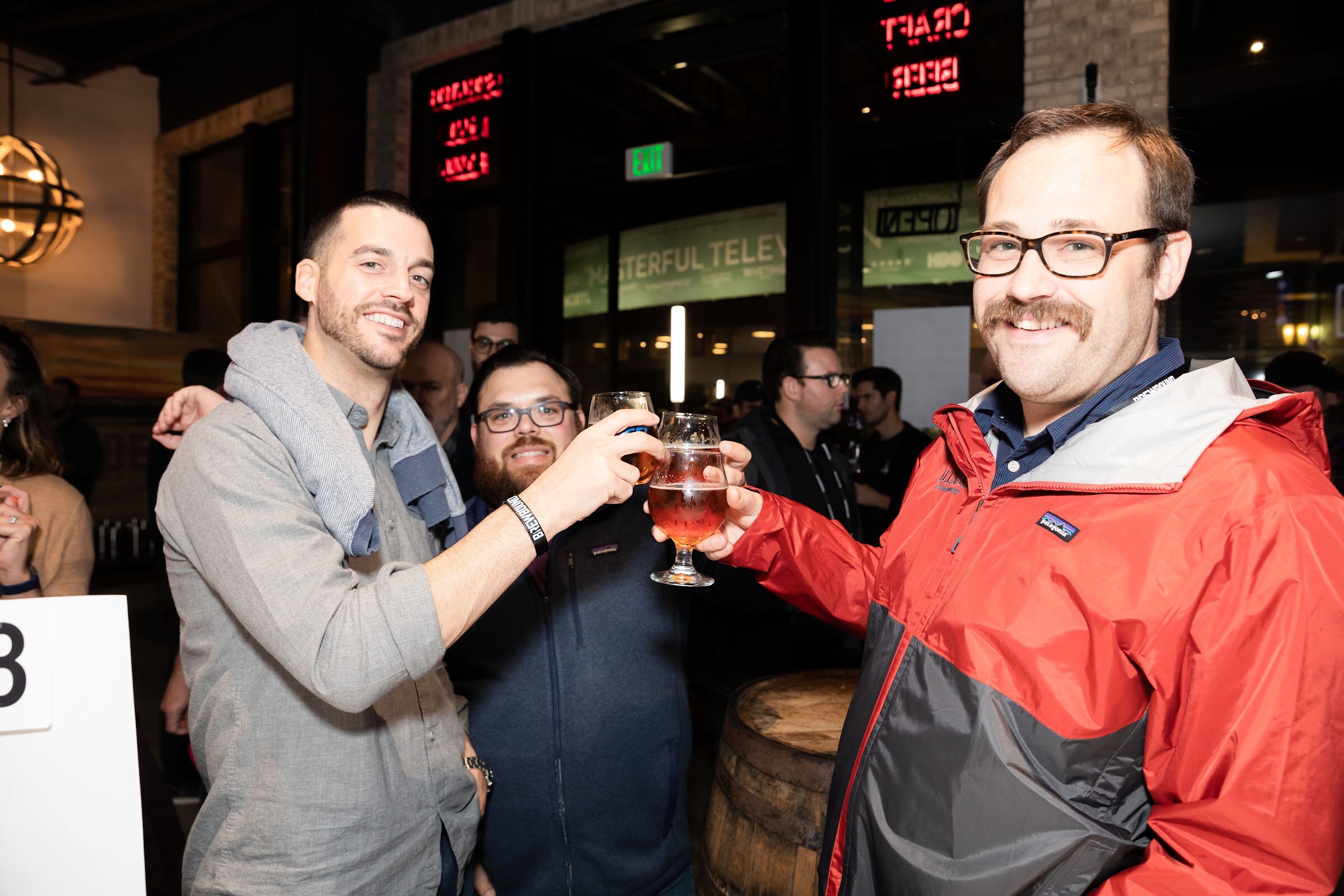 Three men clinking glasses of beer, one in a red and gray jacket.