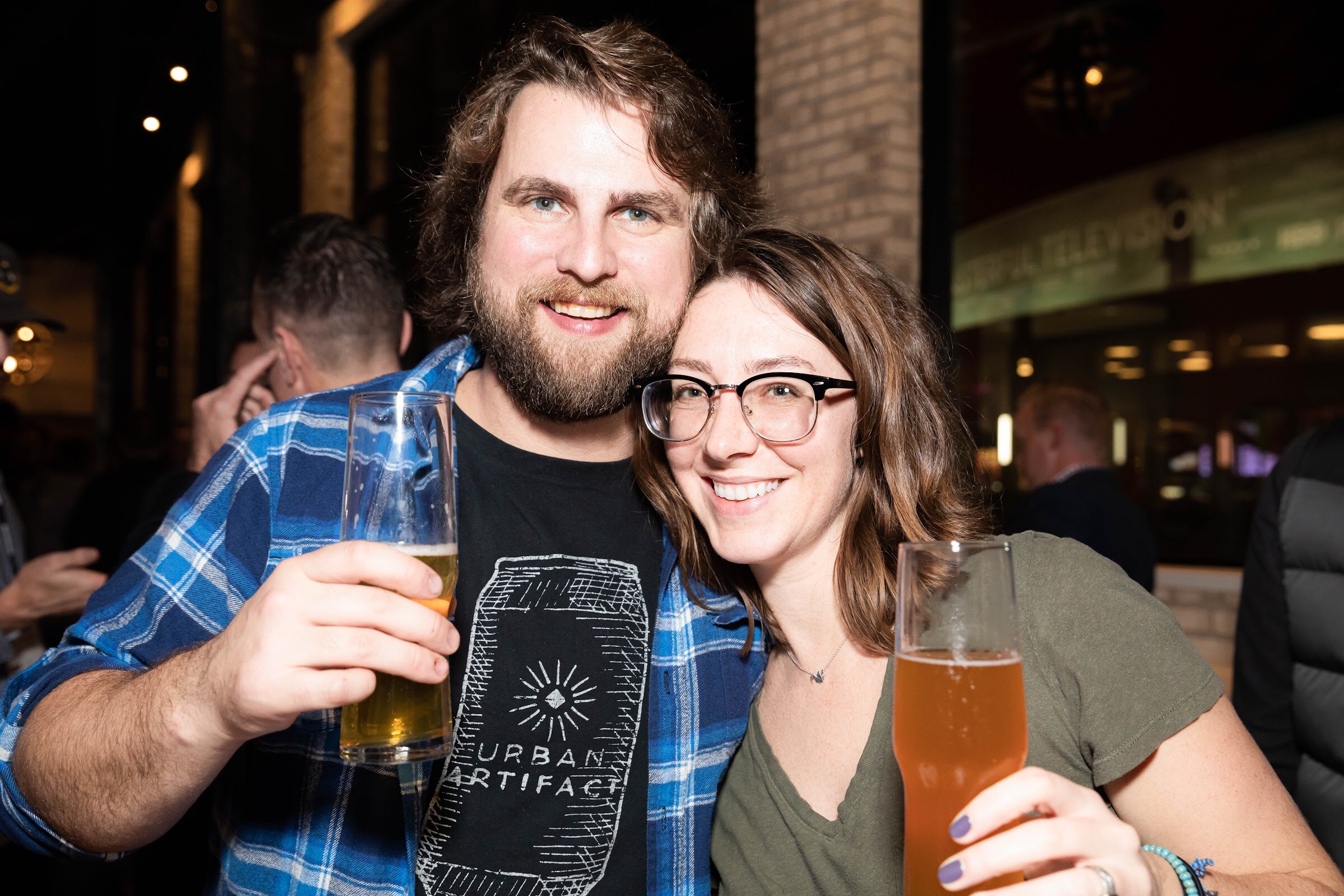 Bearded man in a blue plaid shirt and a woman in glasses holding glasses of beer.