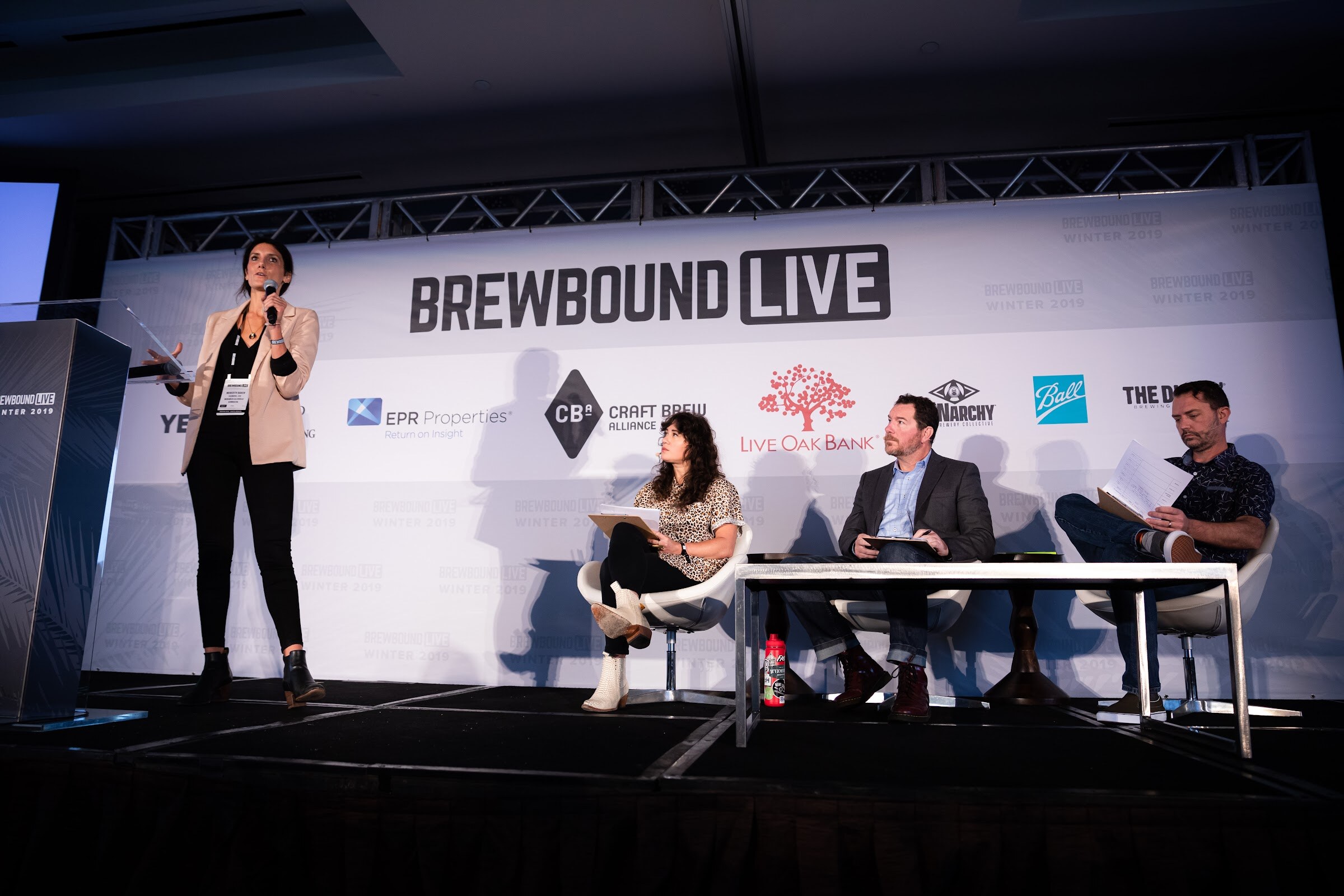 Woman in a blazer at a lectern beside three seated judges on a stage with the Brewbound Live backdrop.