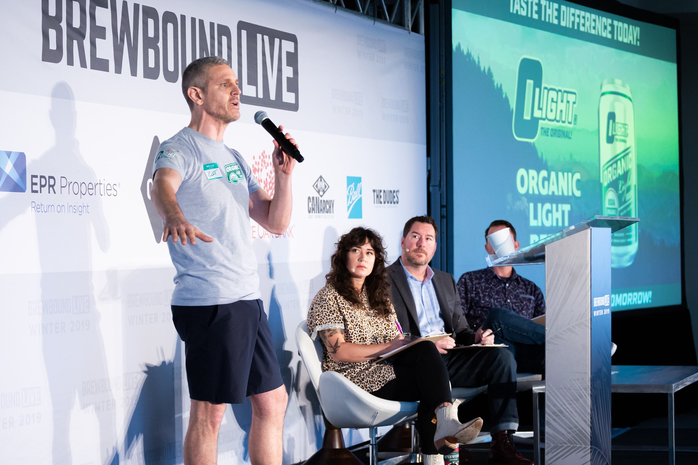 Man in gray shirt and shorts speaking beside a projected Organic Light can while three judges sit behind him.