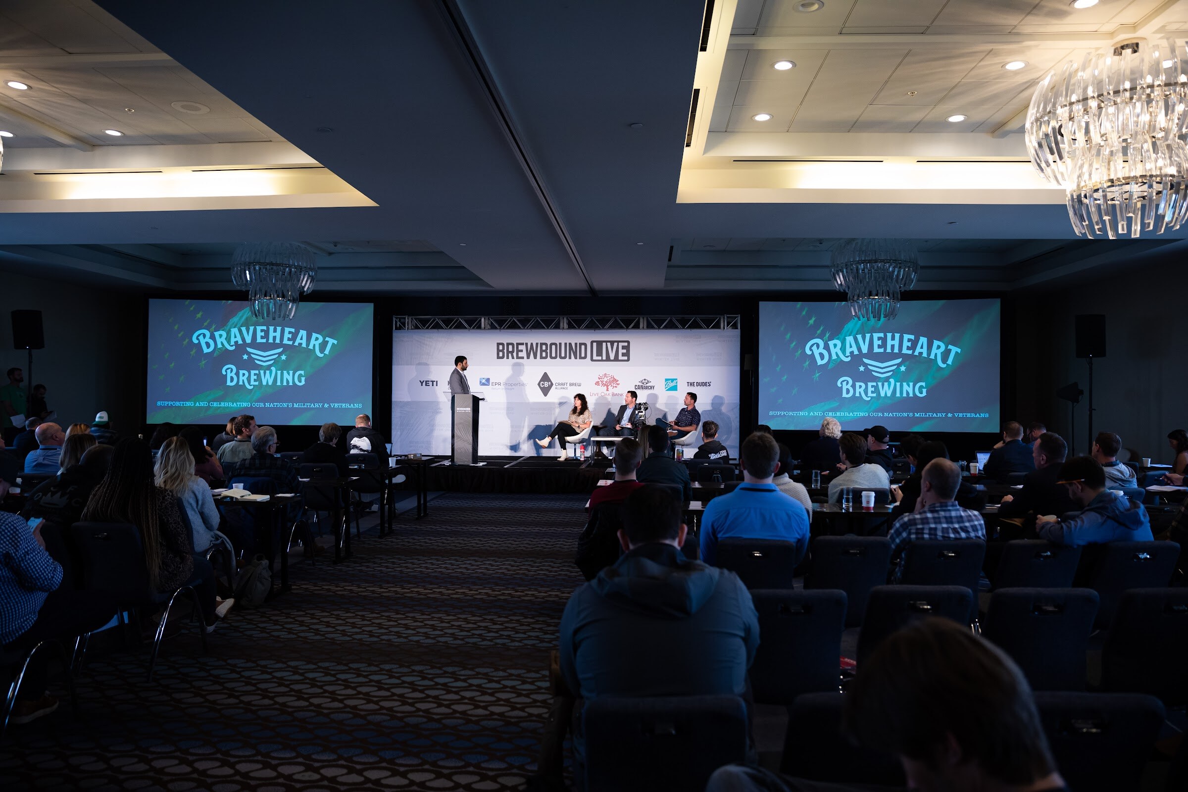 Dark ballroom with audience seated in rows, a stage with judges and two screens showing the Braveheart Brewing logo.