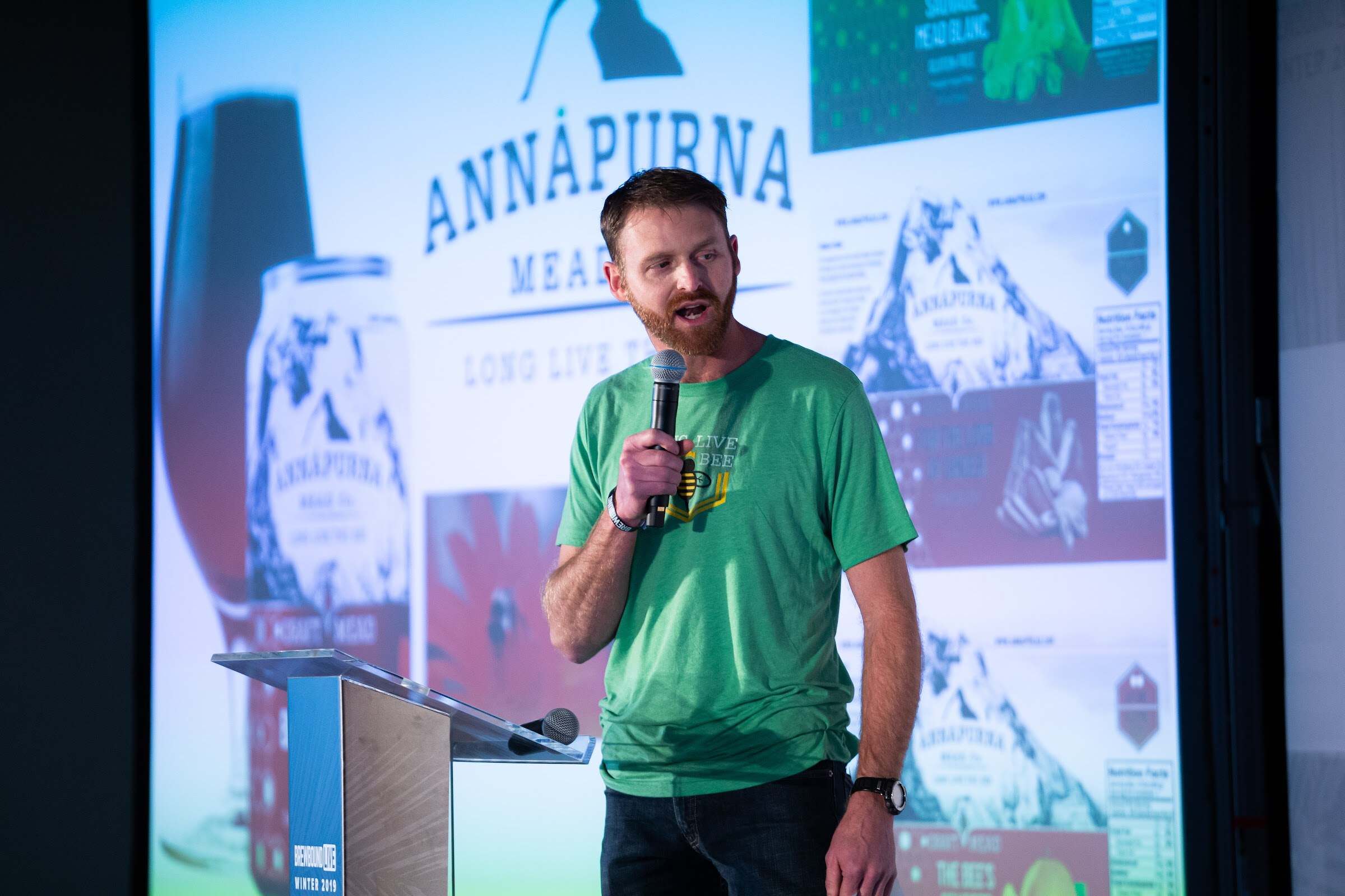 Bearded man in a green T-shirt with a microphone in front of a large screen showing Annapurna Mead can designs.