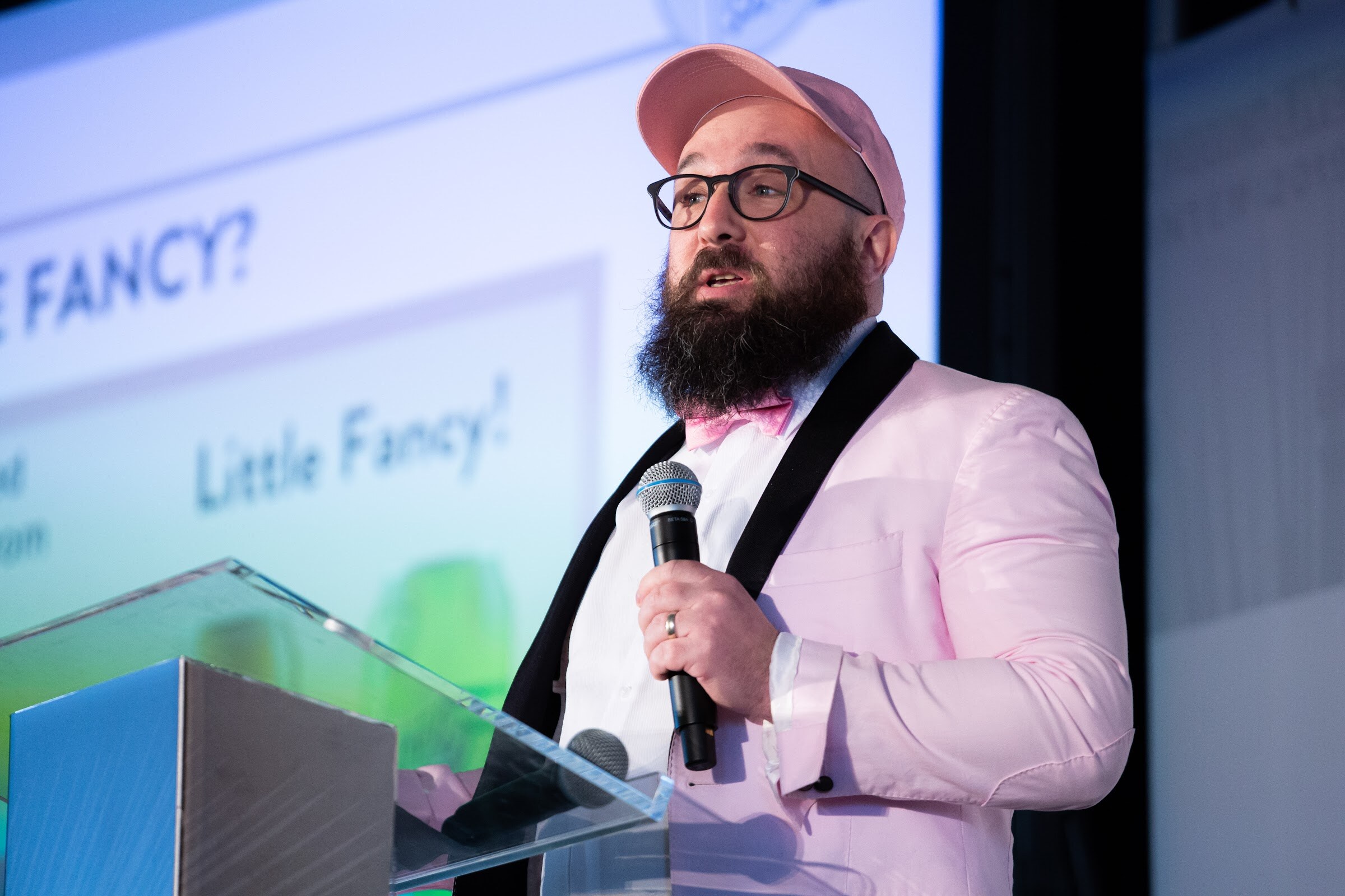 Bearded man in a pink blazer, glasses and bow tie speaking into a microphone in front of a Little Fancy slide.