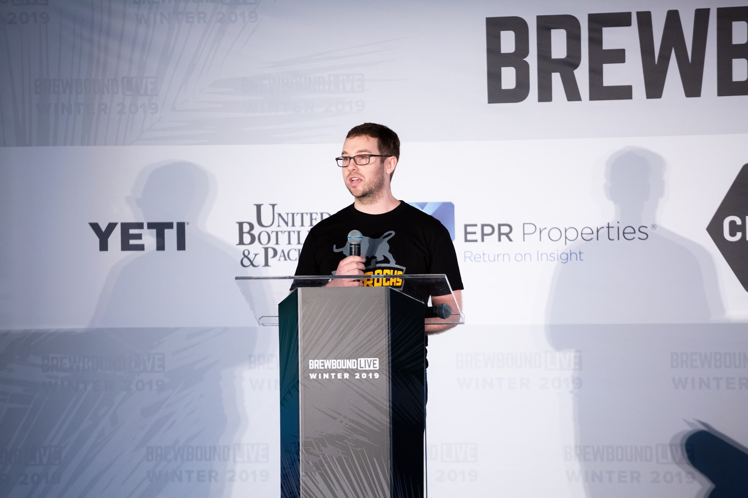 Man in glasses and a black T-shirt at a lectern in front of the Brewbound Live sponsor backdrop.