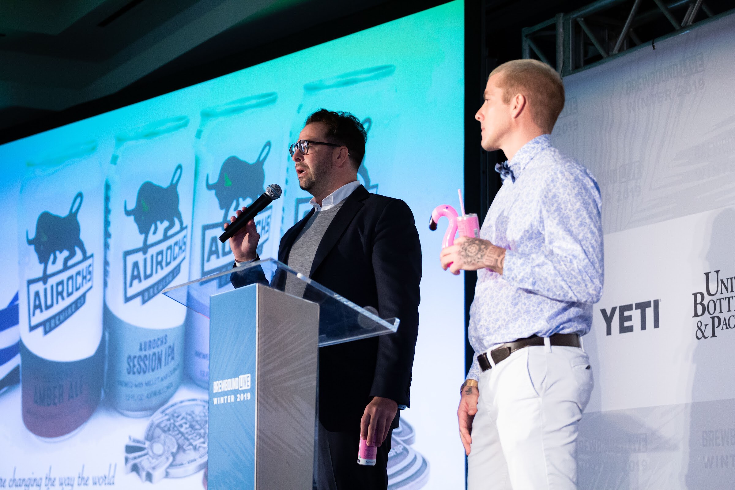 Man in a blazer at a lectern with a microphone beside a man in a floral shirt holding a pink flamingo cup, in front of Aurochs cans on screen.