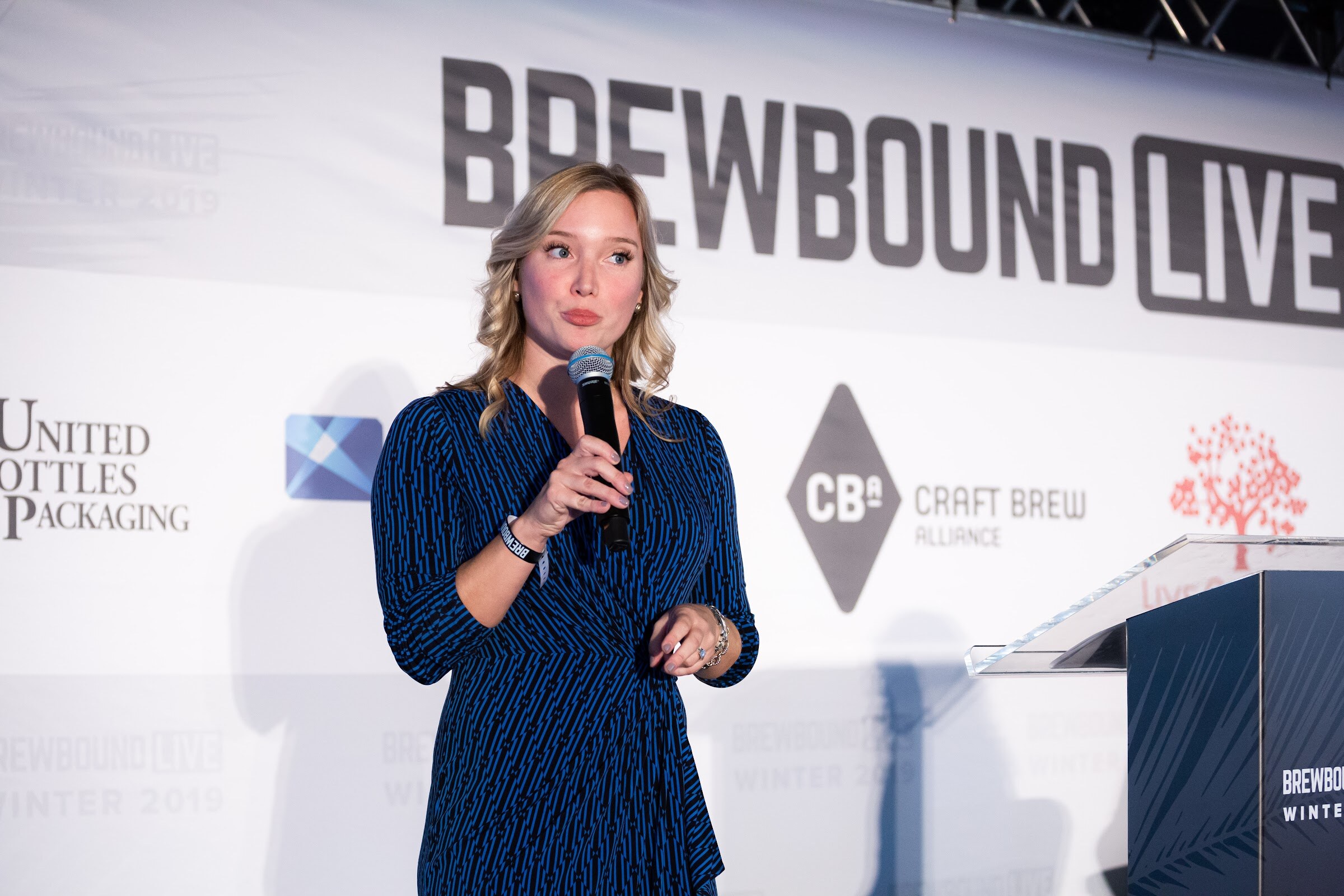 Woman in a navy patterned wrap dress holding a microphone in front of the Brewbound Live backdrop.