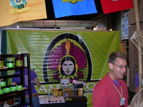 Booth with a large green Sambazon banner showing a face in a feathered headdress. A man in a red shirt and glasses stands at right, and products sit on shelves at left.