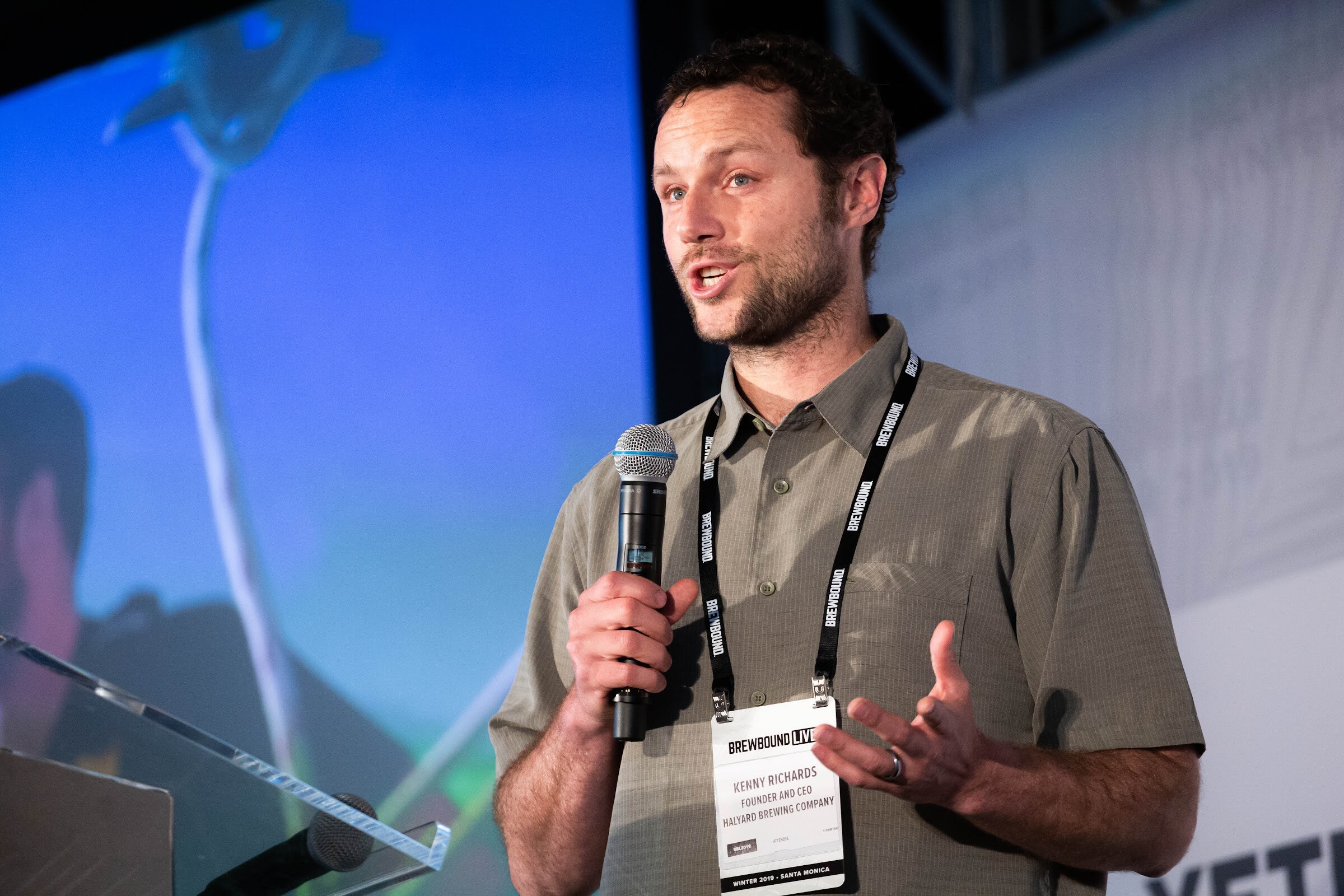 Bearded man in an olive button-up shirt with a badge holding a microphone and gesturing.
