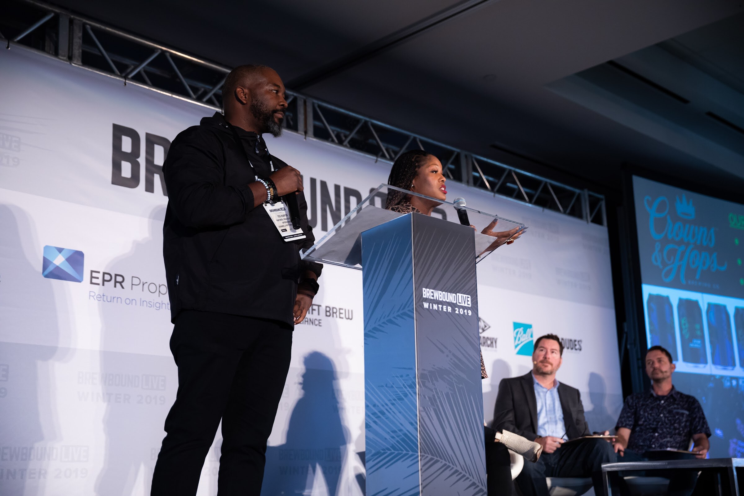Man in black and a woman at a glass lectern with judges seated at right and a Crowns & Hops slide on the side screen.