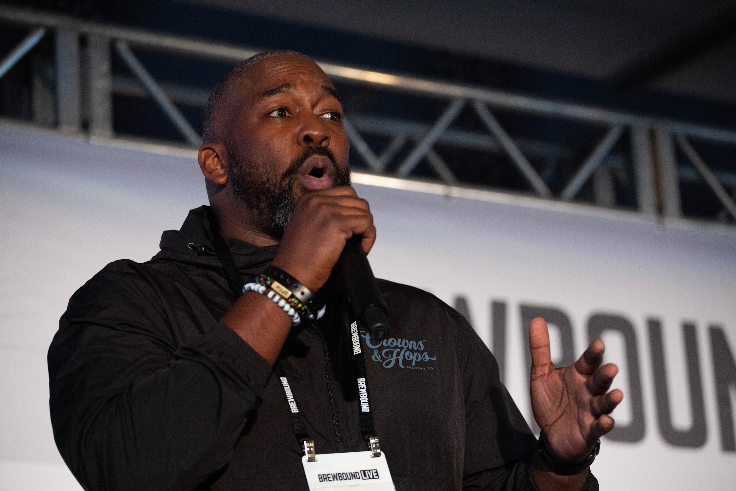 Bearded man in a black Crowns & Hops hoodie with a conference badge speaking into a microphone.