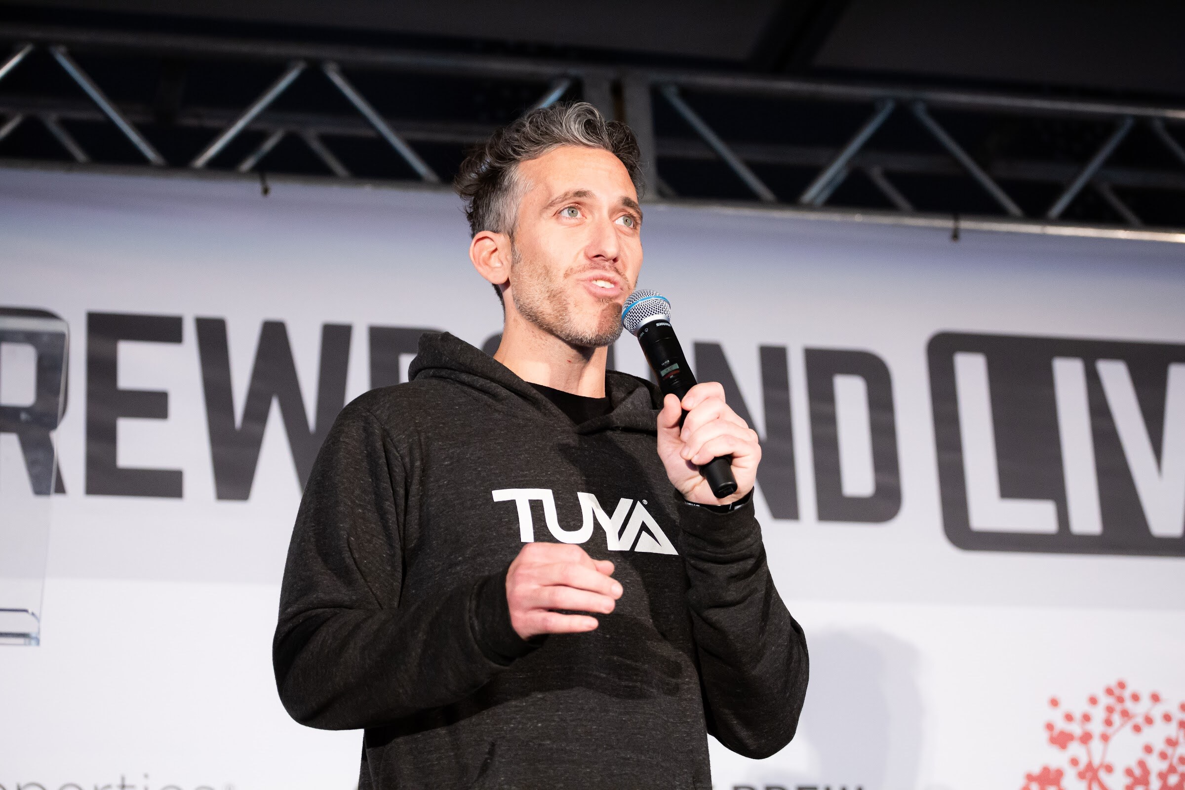 Man with gray-streaked hair in a black TUYA hoodie holding a microphone in front of the Brewbound Live backdrop.