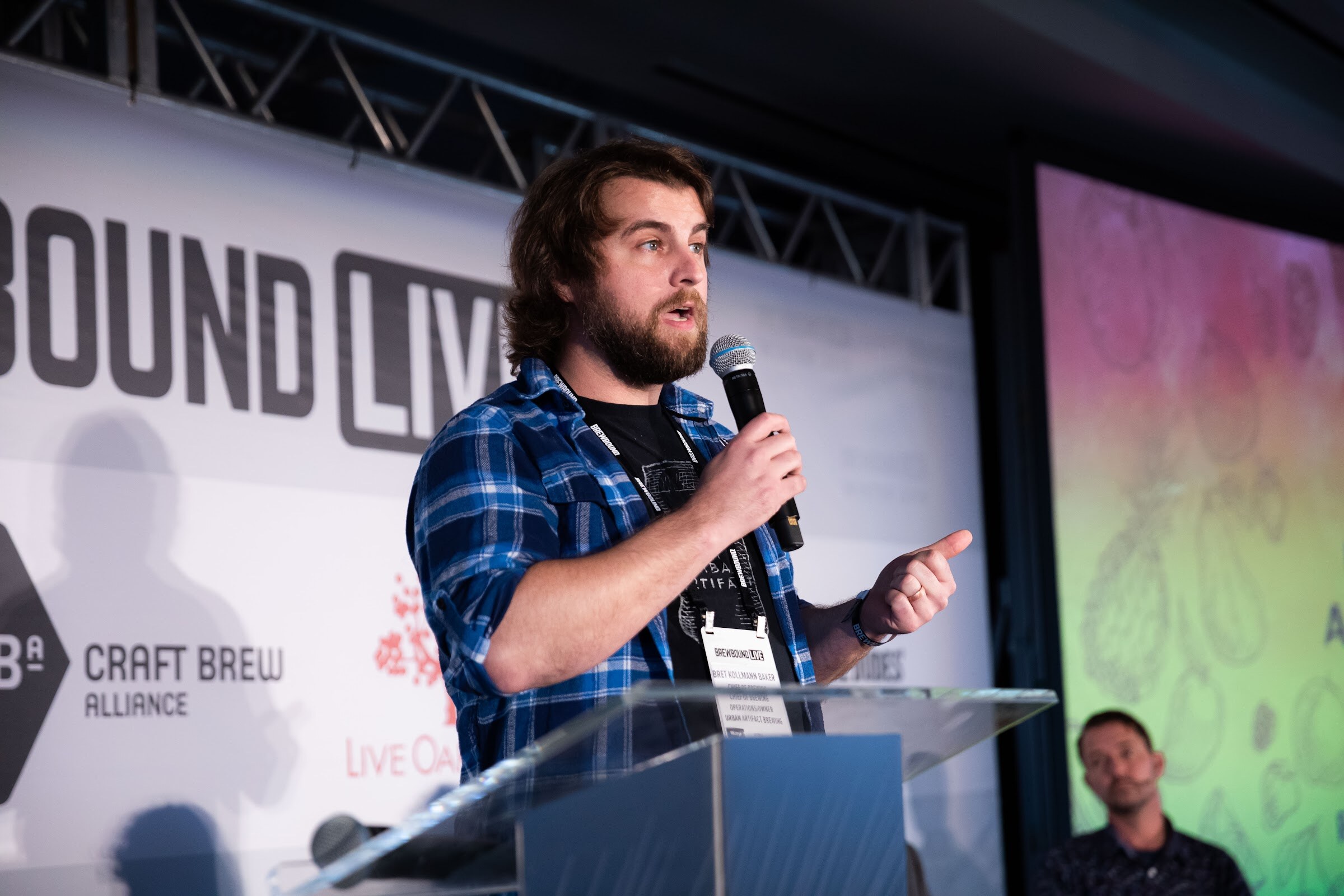 Bearded man in a blue plaid shirt with a badge speaking into a microphone in front of a green and pink slide.
