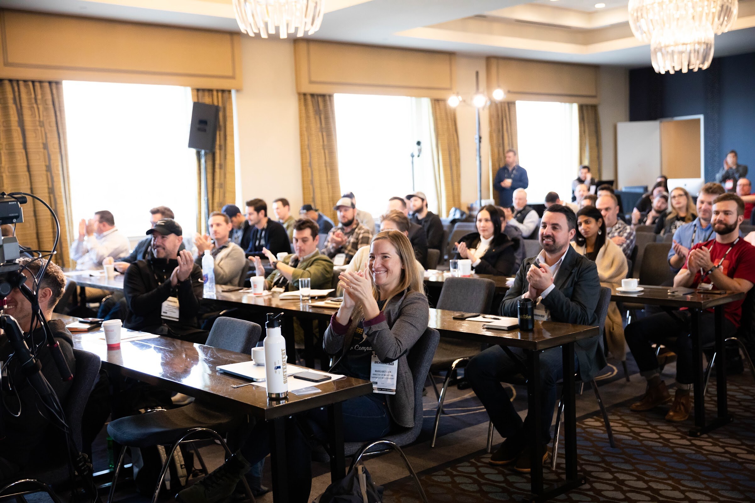 Attendees at long tables in a sunlit ballroom applauding, with a camera operator at left.