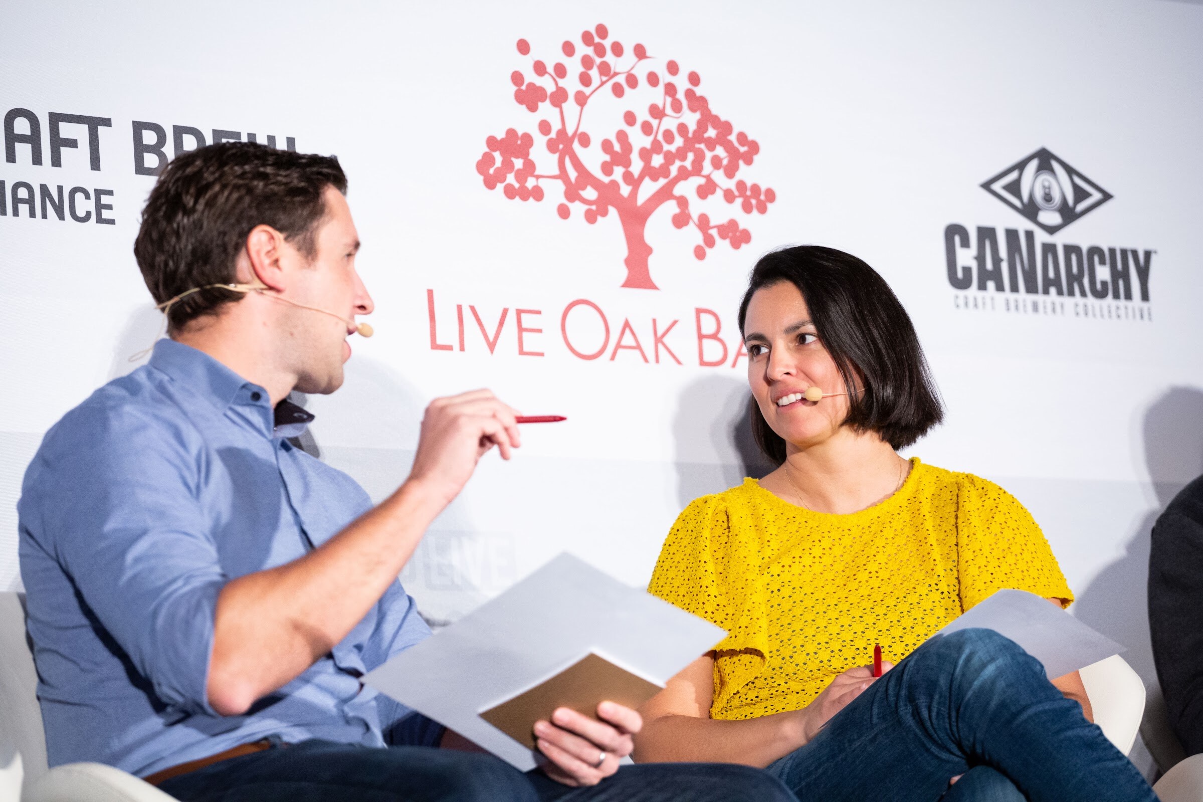 Man in a blue shirt holding a pen and papers talking with a smiling woman in a yellow top.
