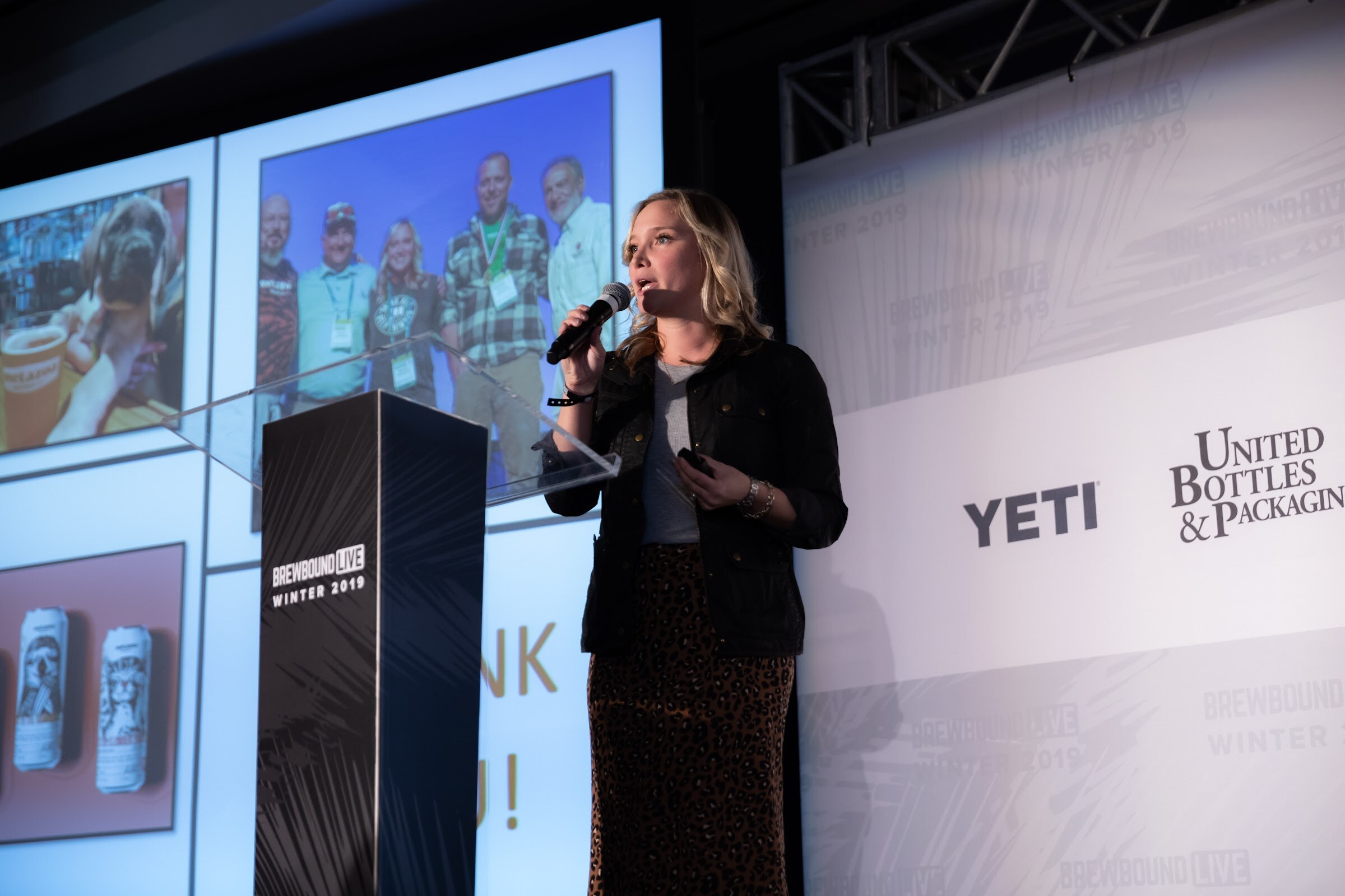 Woman in a black jacket speaking into a microphone at a lectern beside a screen of beer photos.