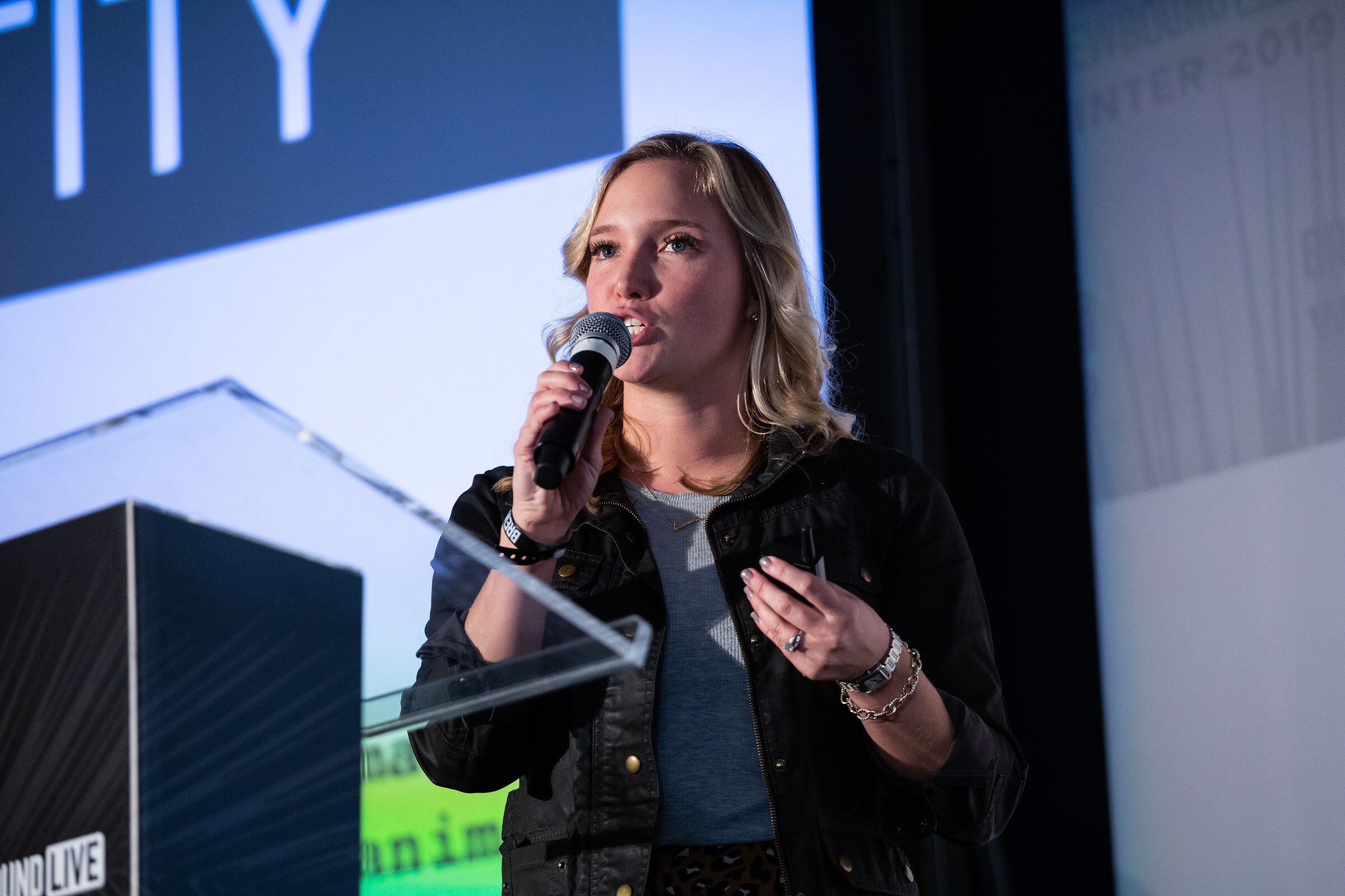 Blonde woman in a black jacket speaking into a microphone in front of a dark screen.
