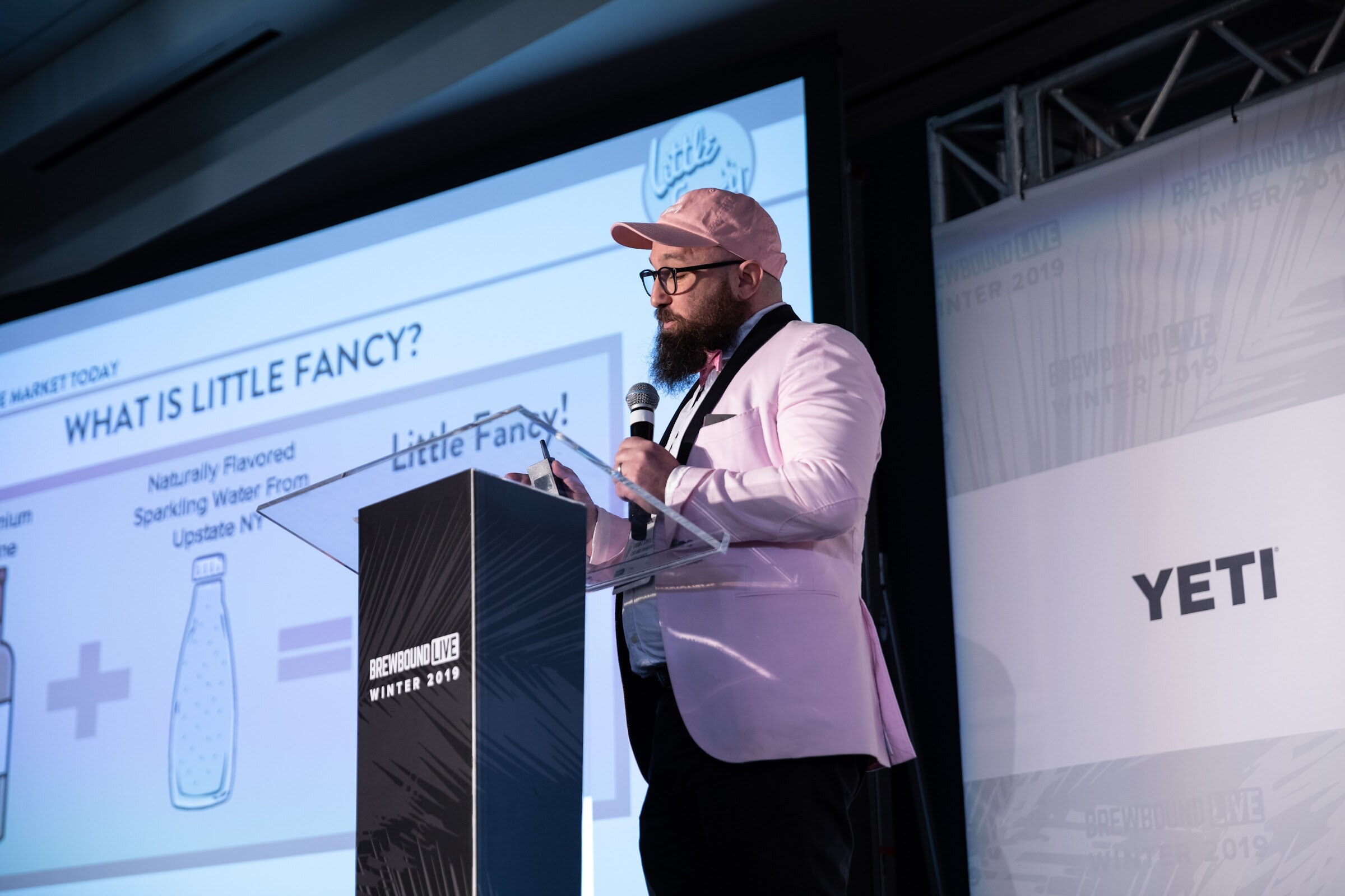 Bearded man in a pink blazer and cap at a lectern beside a screen reading What is Little Fancy.