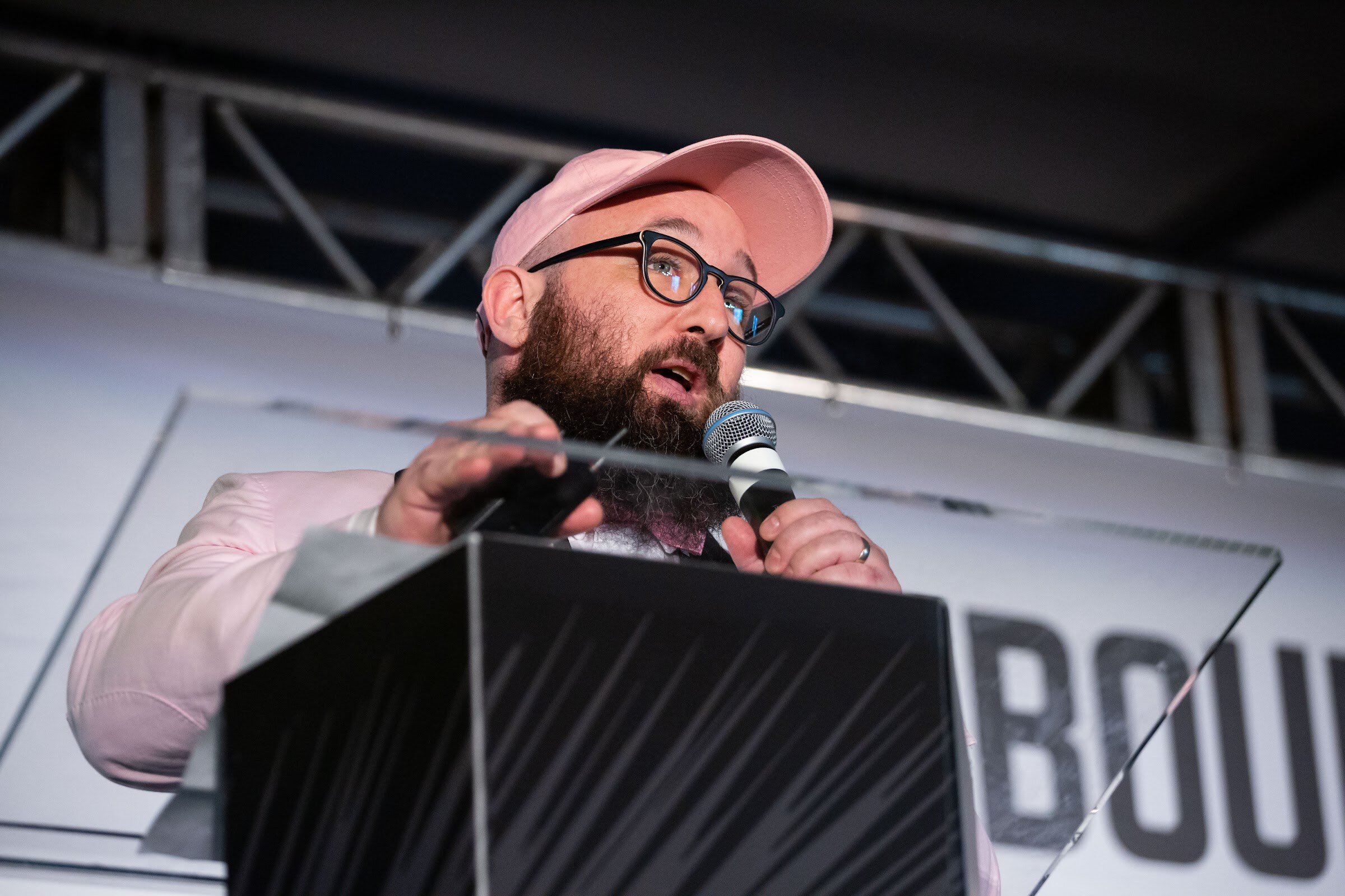 Man with a beard and glasses in a pink cap and blazer seen from below at a black lectern.