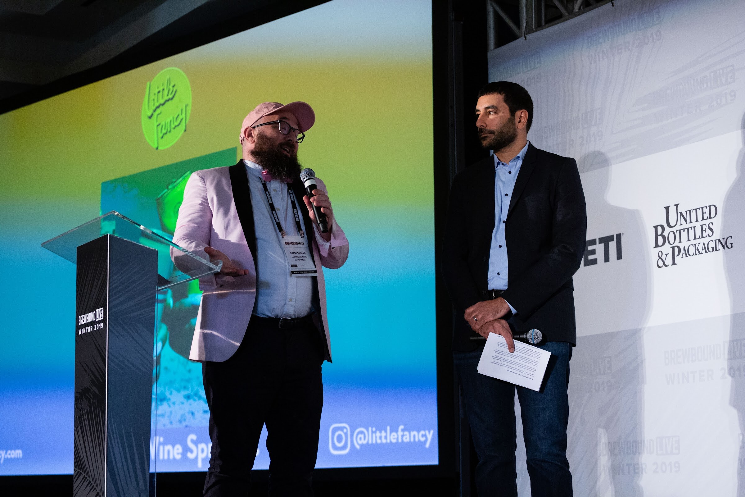 Man in a pink blazer at a lectern beside a man in a dark blazer, in front of a Little Fancy screen.