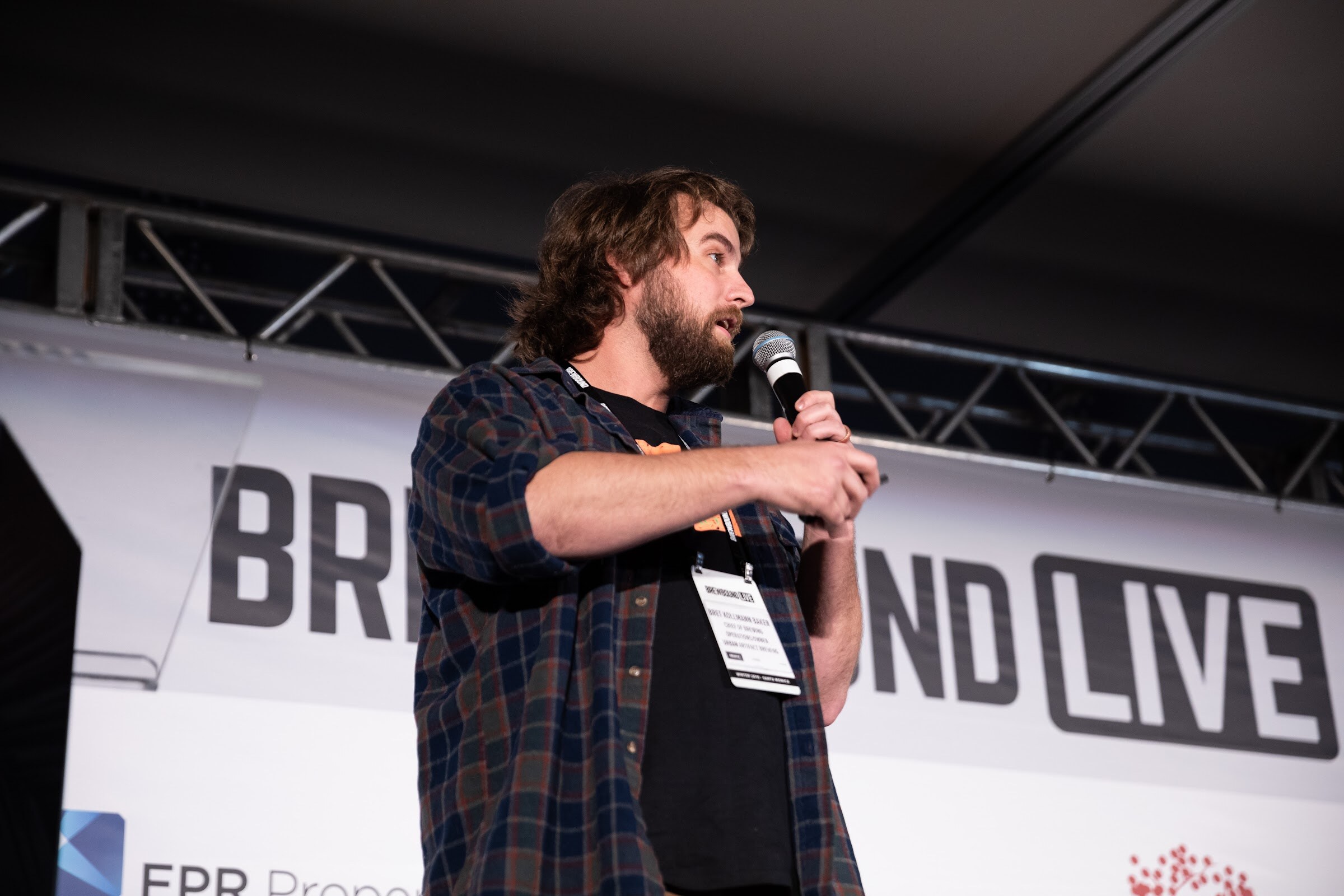 Bearded man in a blue and brown plaid shirt with a badge speaking into a microphone.
