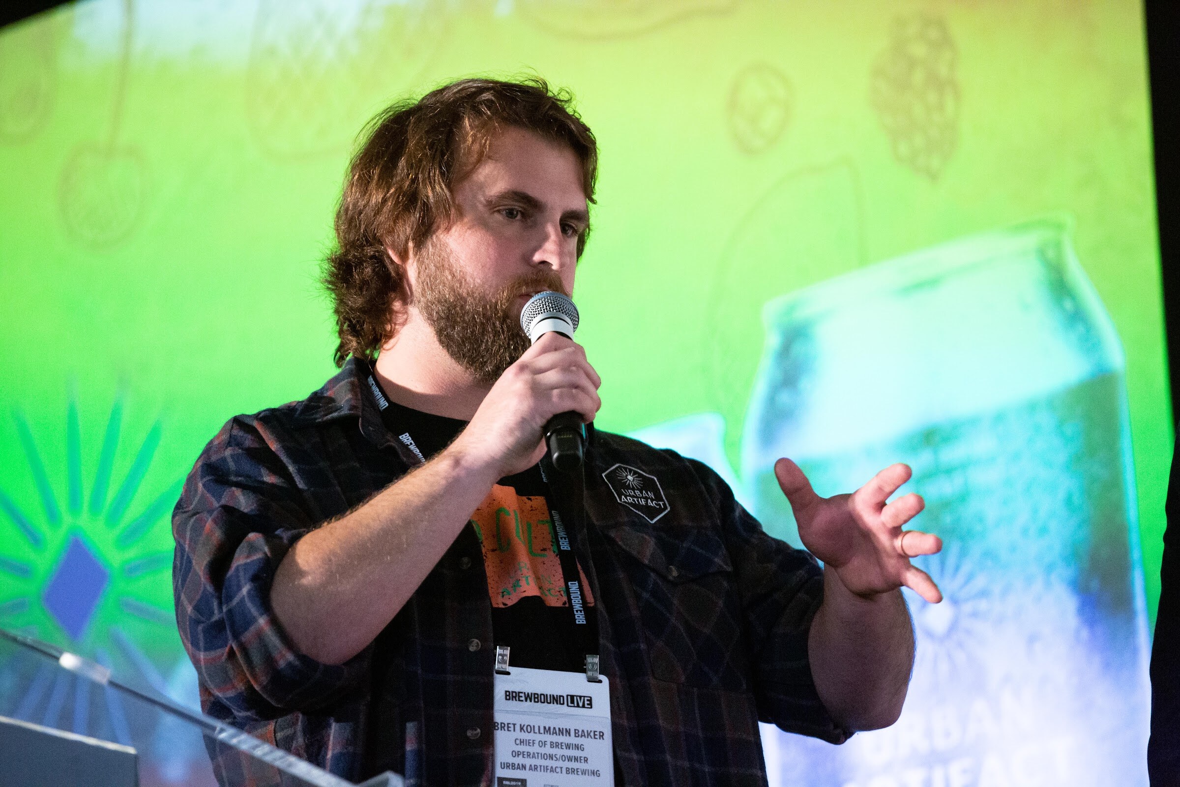 Bearded man in a plaid shirt with a conference badge speaking into a microphone before a green projected can.