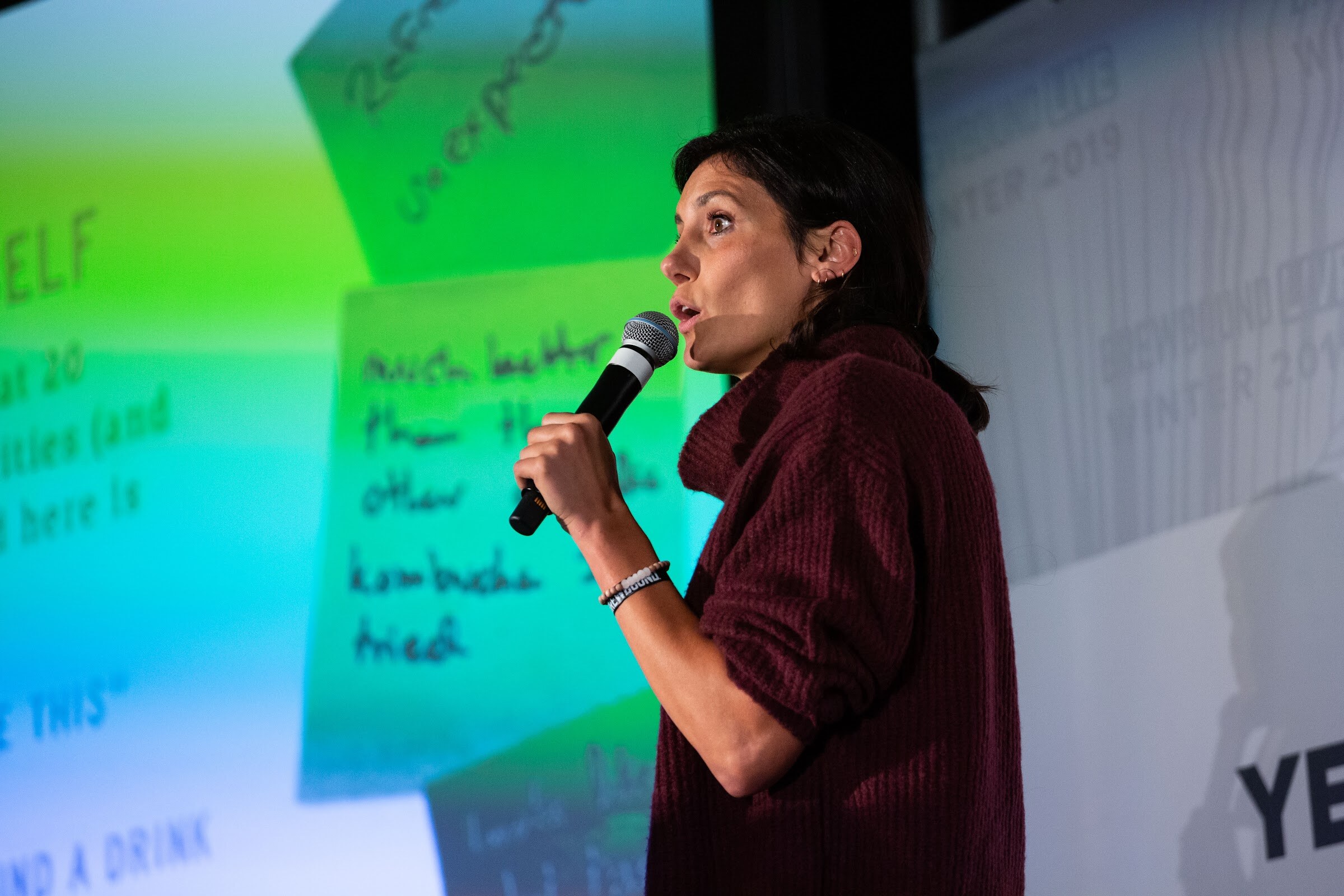 Woman in a maroon sweater speaking into a microphone beside a slide of green handwritten sticky notes.