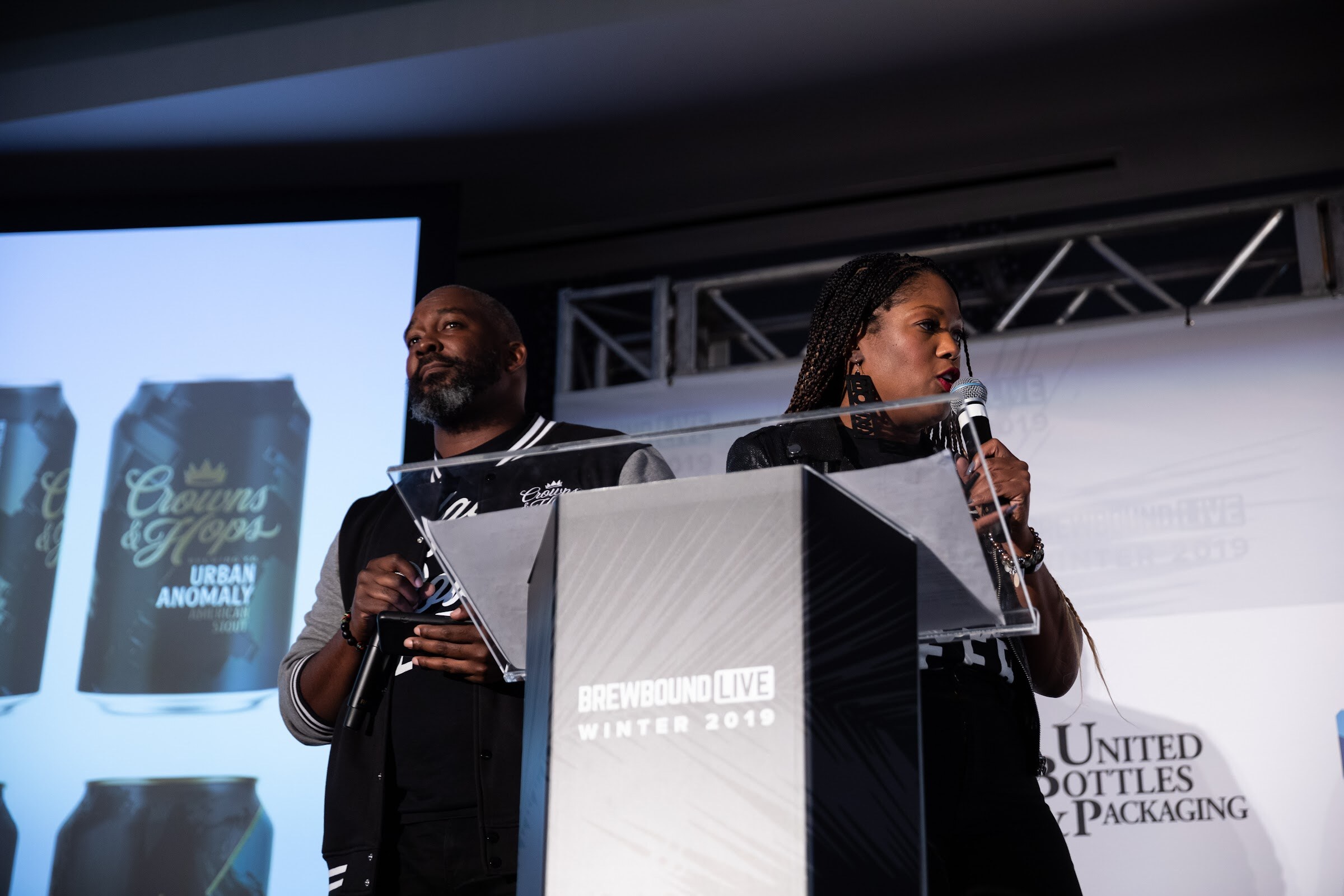 Man and woman at a glass lectern in front of a screen showing Crowns & Hops Urban Anomaly cans.