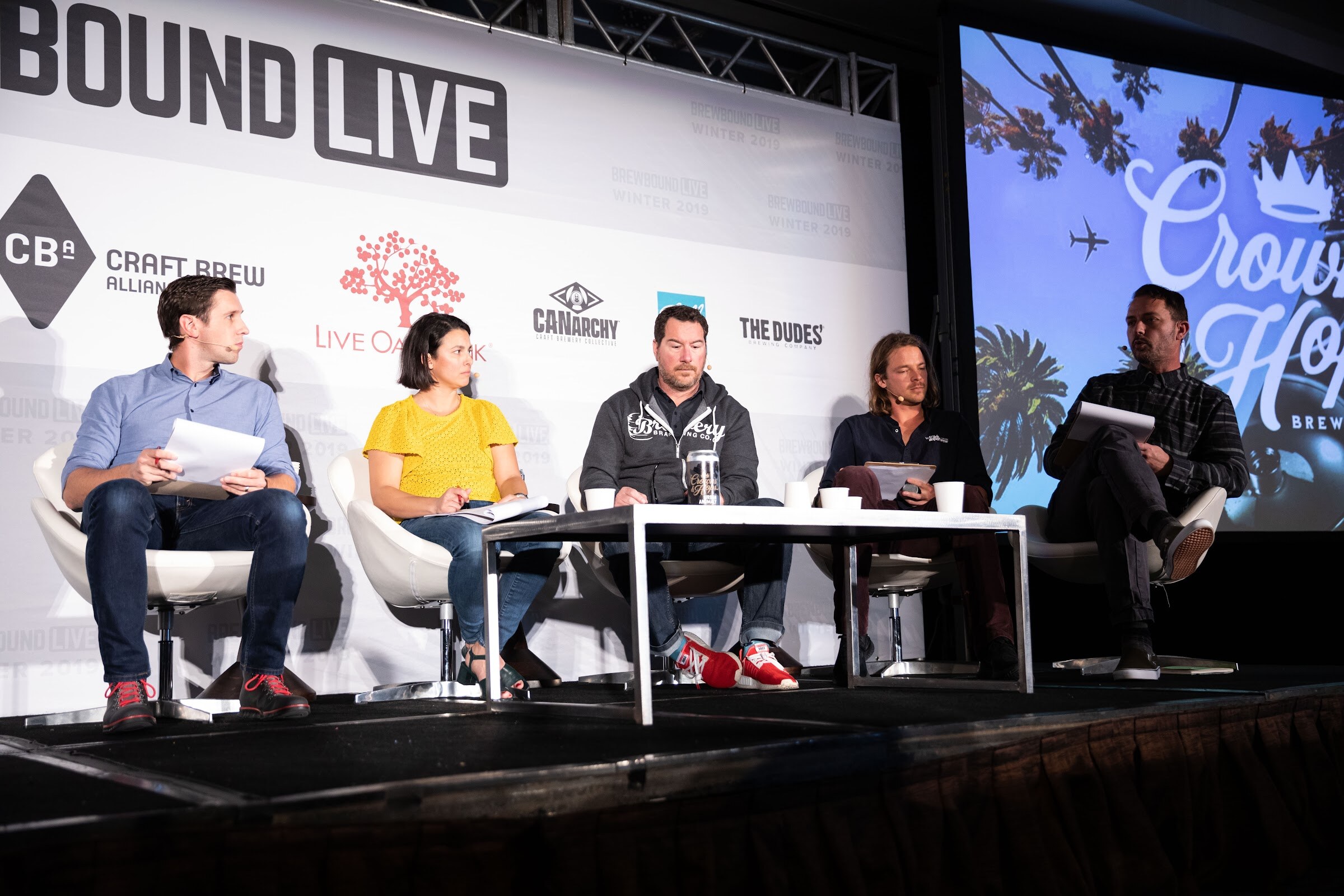 Five judges seated in white chairs on stage next to a screen with the Crowns & Hops logo over palm trees.