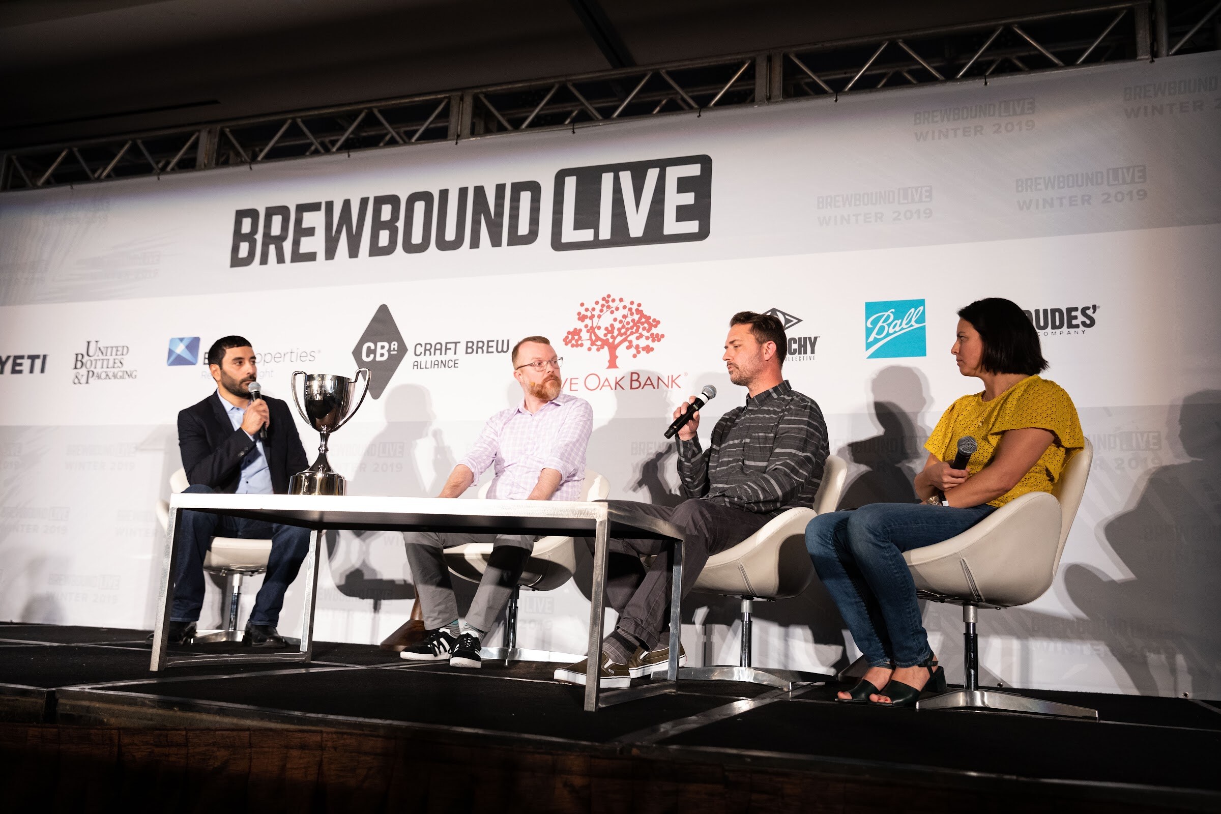 Four people seated on stage in front of the Brewbound Live backdrop with a silver trophy on the table.