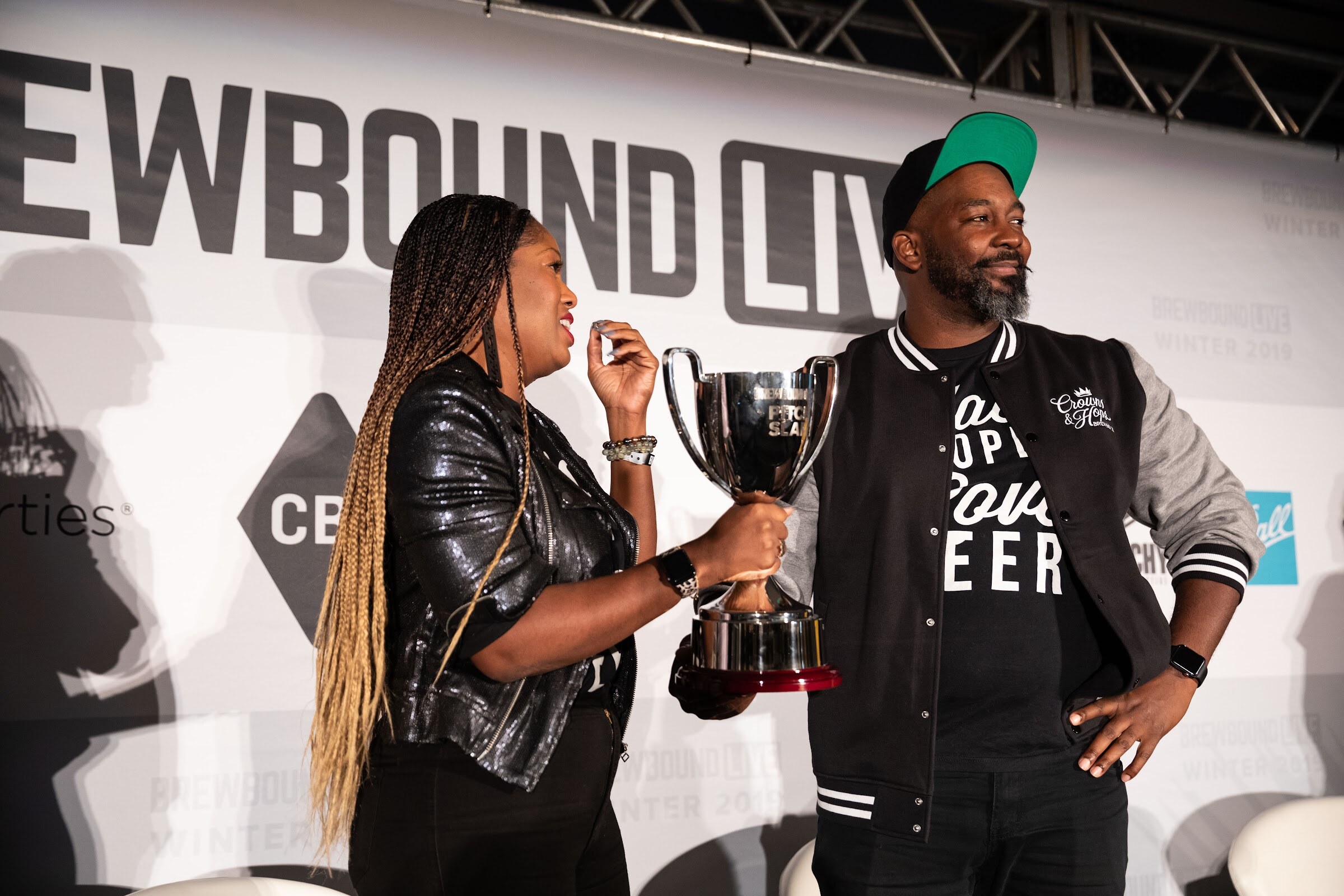 Woman in a black leather jacket holding a silver cup next to a man in a Crowns & Hops varsity jacket and green cap.