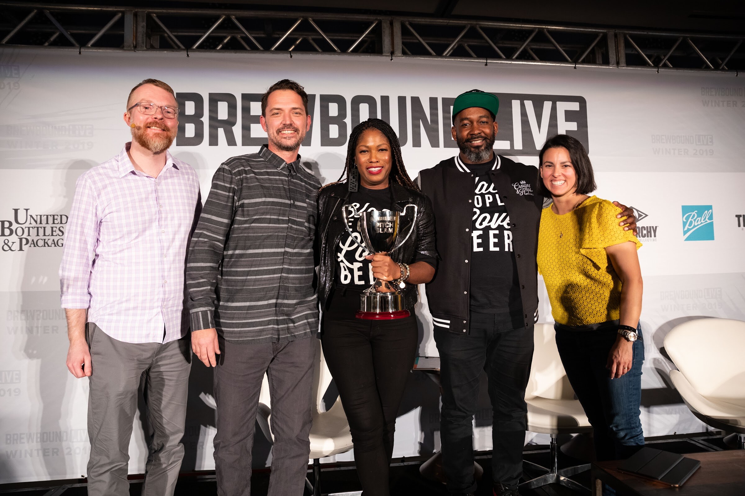 Five people standing in a row on stage, the center two holding a silver trophy in Black People Love Beer shirts.