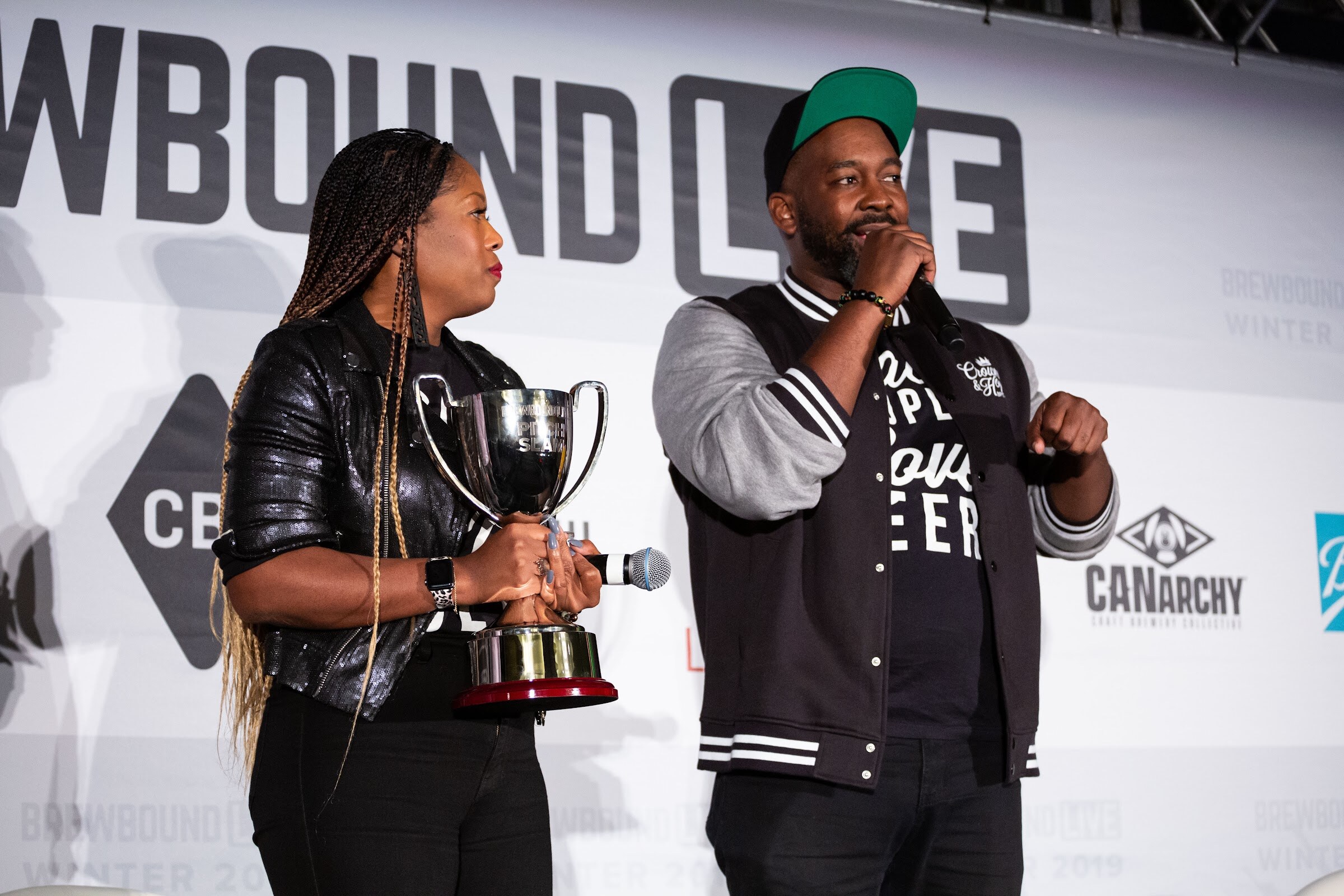 Woman holding a silver trophy next to a man in a varsity jacket speaking into a microphone.