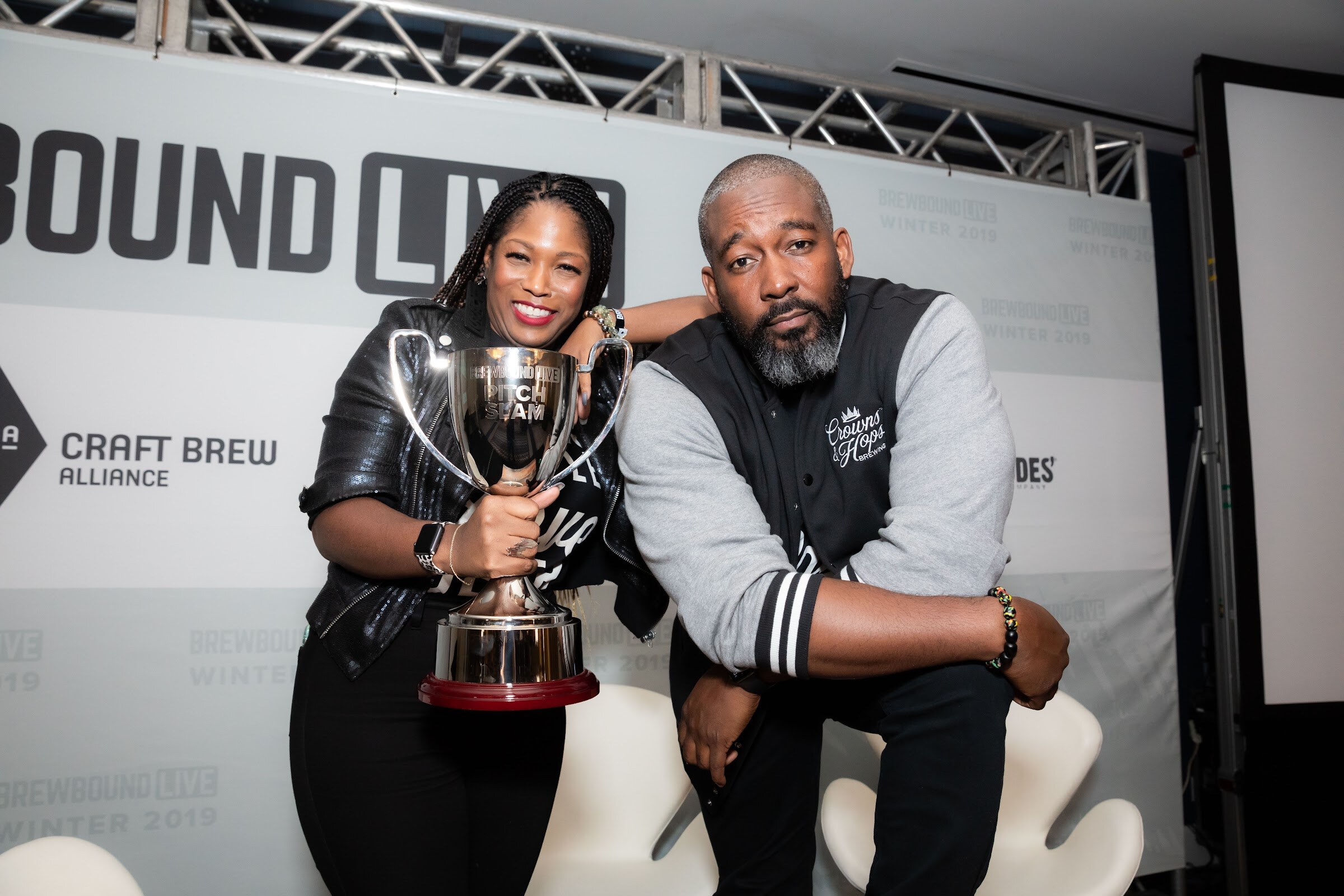 Woman in a leather jacket holding a silver trophy leaning against a man in a gray and black varsity jacket.