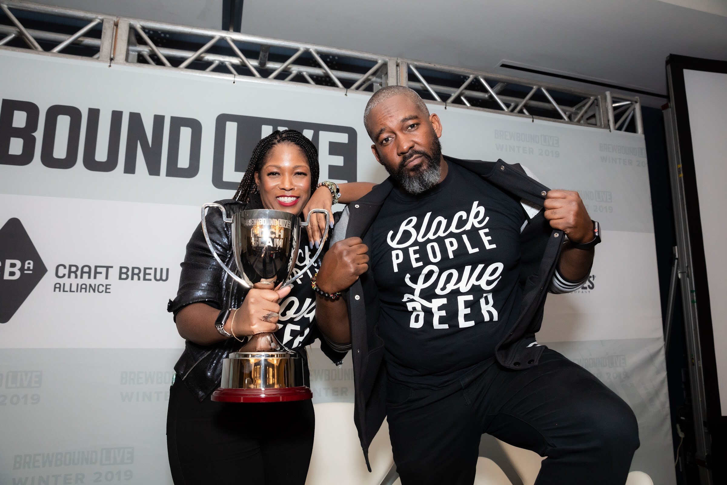 Man opening his jacket to show a T-shirt reading Black People Love Beer beside a woman holding a silver trophy.