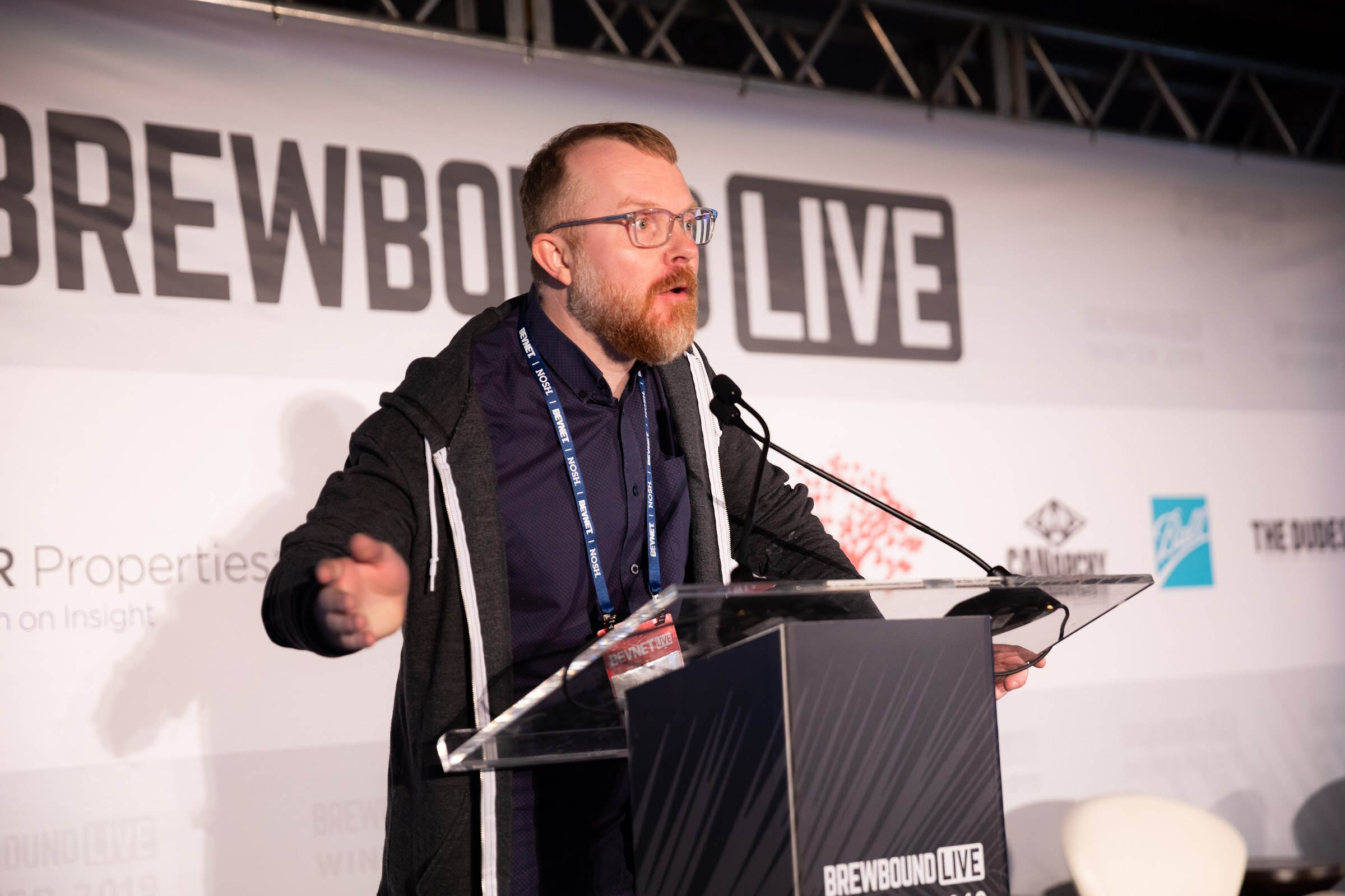 Man with a beard in a dark jacket speaking at a lectern in front of the Brewbound Live backdrop.